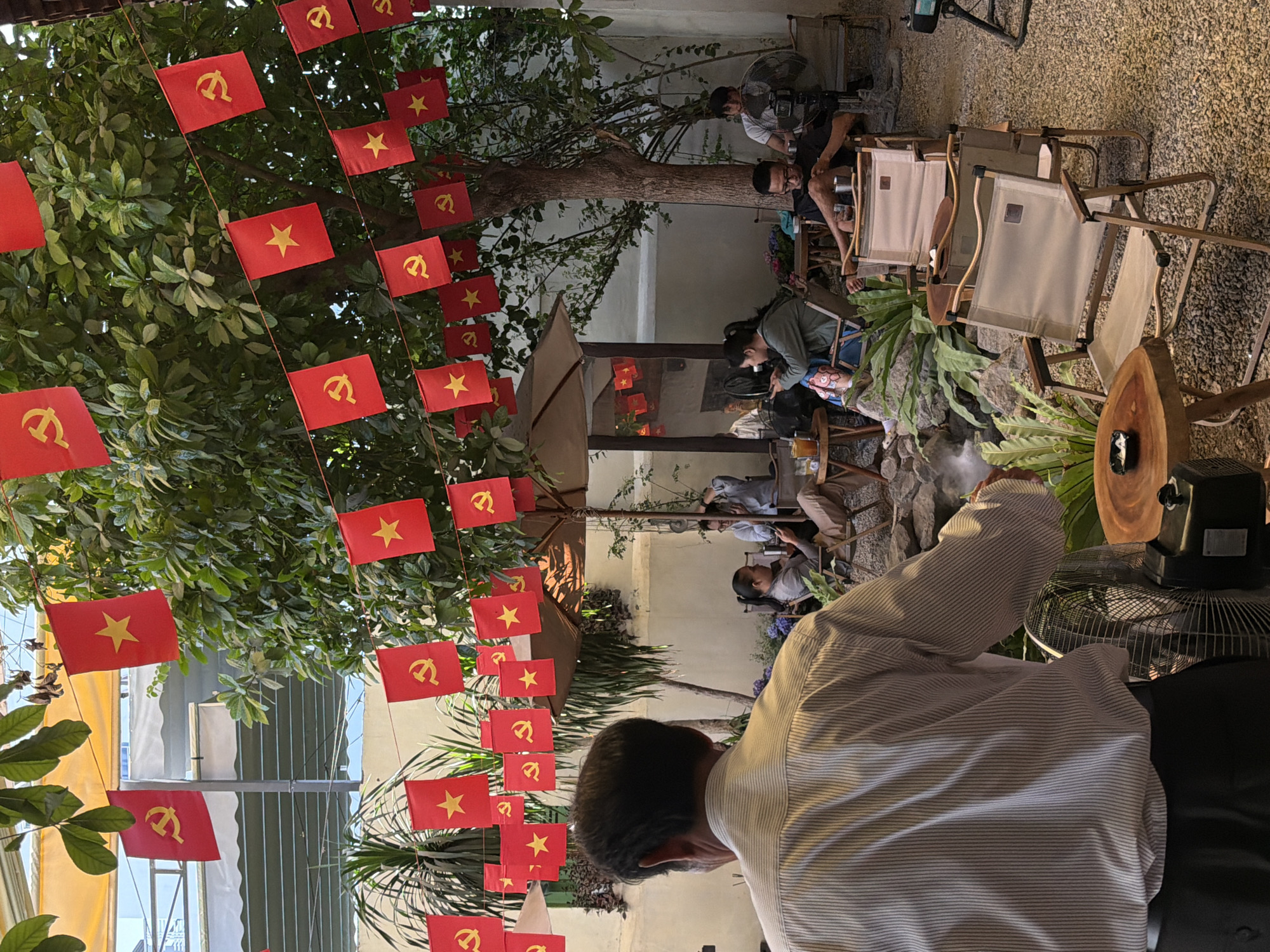 The image depicts an outdoor cafe located in Bac Nha Trang Ward, Vietnam. The cafe is decorated with numerous small red flags featuring a yellow star and hammer and sickle emblem, strung along wires overhead and draped from trees. In the foreground, the back of a man in a striped shirt is visible, facing away from the camera. Behind him, several people are seated at tables, some engaged with their phones. The cafe features a gravel ground, an assortment of chairs, and tables. Lush greenery, including trees and plants, surrounds the seating area. The background reveals a light-colored building structure and a partially visible awning.