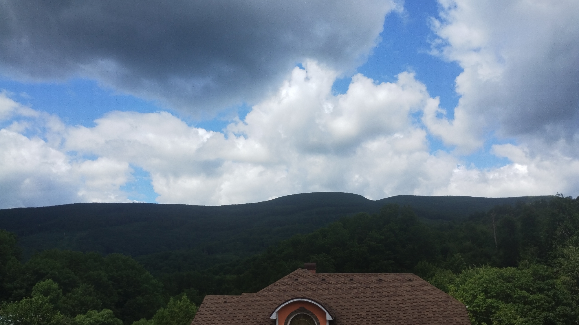 A rooftop of a building with brown shingles and a partial orange window is visible in the foreground, with lush green trees surrounding it. Beyond the trees, a dark, forested mountain range stretches across the horizon under a sky filled with dramatic clouds, featuring both dark storm clouds and bright blue patches. The scene appears to be a landscape view, possibly from a residential area in Syniak, Ukraine, given the presence of the building and the natural mountainous terrain.