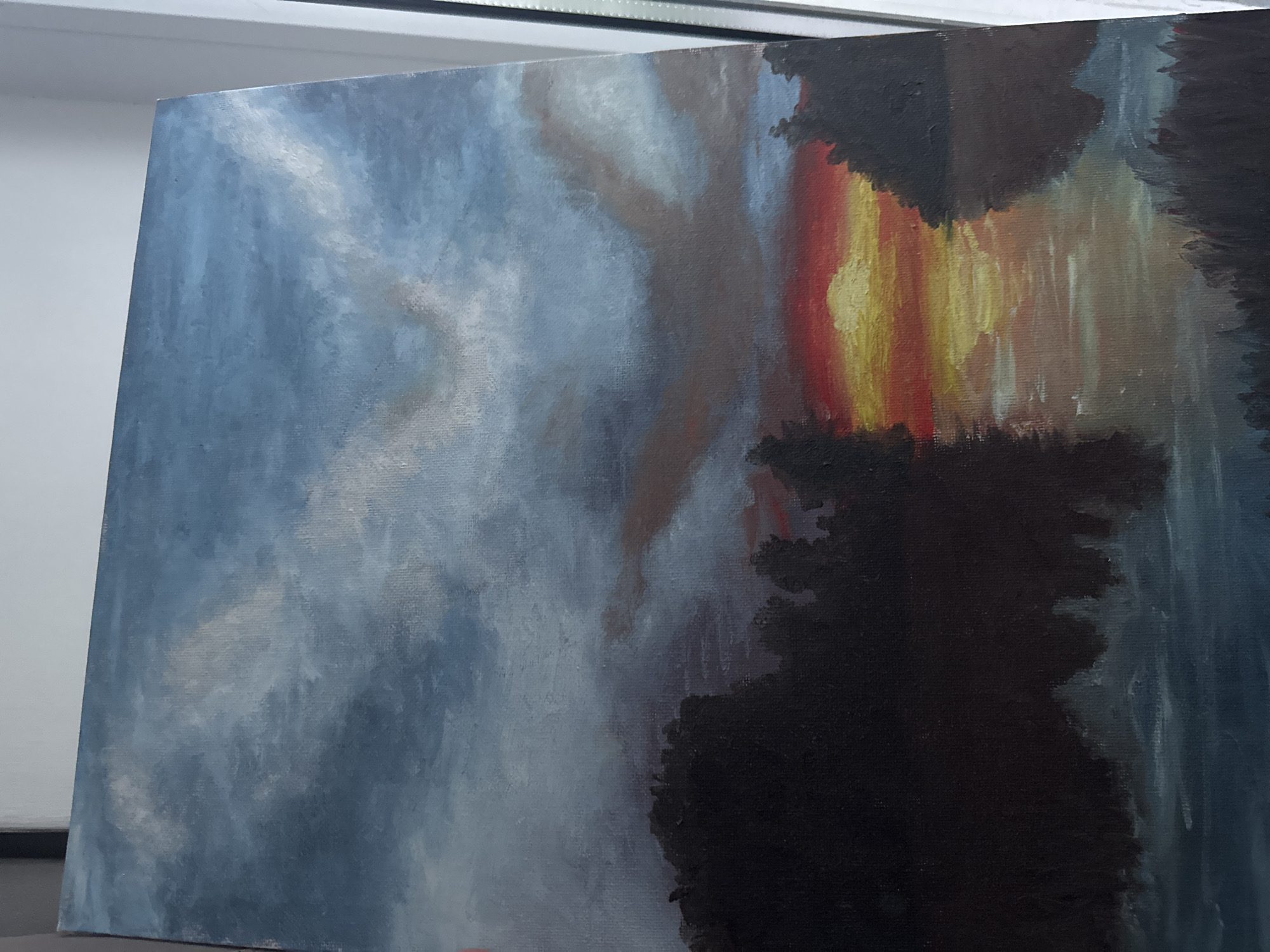 This is a close-up, angled photograph of an oil painting on canvas, which is placed on an easel. The painting depicts a stormy or twilight sky with dark, silhouetted shapes of trees or landmasses in the foreground and midground. The sky is rendered in shades of blue, grey, and white, with swirling cloud patterns. A dramatic sunrise or sunset is visible on the right side of the painting, featuring intense bands of yellow, orange, and red. The dark shapes in the foreground are almost black, with some textured brushstrokes. The overall mood of the painting is dramatic and somewhat somber, possibly evoking a sense of awe or foreboding. There is no visible text in the image. The lighting suggests the photograph was taken indoors, possibly in a studio or gallery setting, with bright ambient light coming from above and to the left. The painting itself appears to be in progress, as evidenced by the visible texture of the canvas and the brushstrokes.