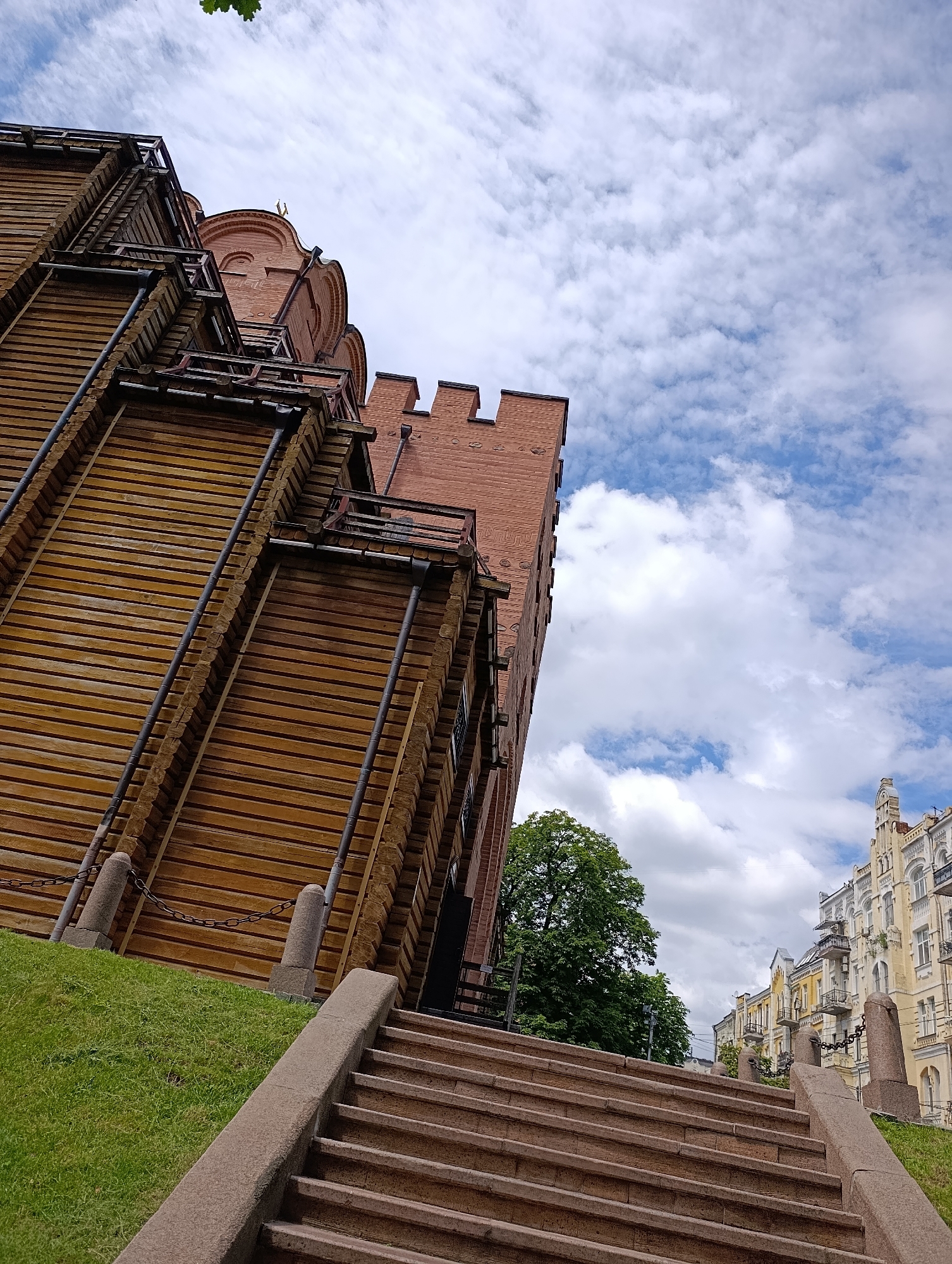 The image shows a low-angle view of the Golden Gate in Kyiv, Ukraine, against a cloudy blue sky. The gate is a historical monument, a reconstruction of a medieval city gate. In the foreground, a wide stone staircase ascends, leading towards the gate. The gate itself is a prominent structure, featuring a large wooden facade with dark metal accents and a red brick upper section topped with a golden dome. To the right of the gate, a lush green lawn slopes upwards, bordered by a low stone wall and a chain fence. Further to the right, beyond the gate, several multi-story buildings with distinctive architecture, characteristic of Kyiv's historic streets, line a road. A large, mature tree with dense green foliage stands between the gate and the buildings. The lighting suggests it is daytime, with diffused light from the overcast sky. There are no people visible in the image.