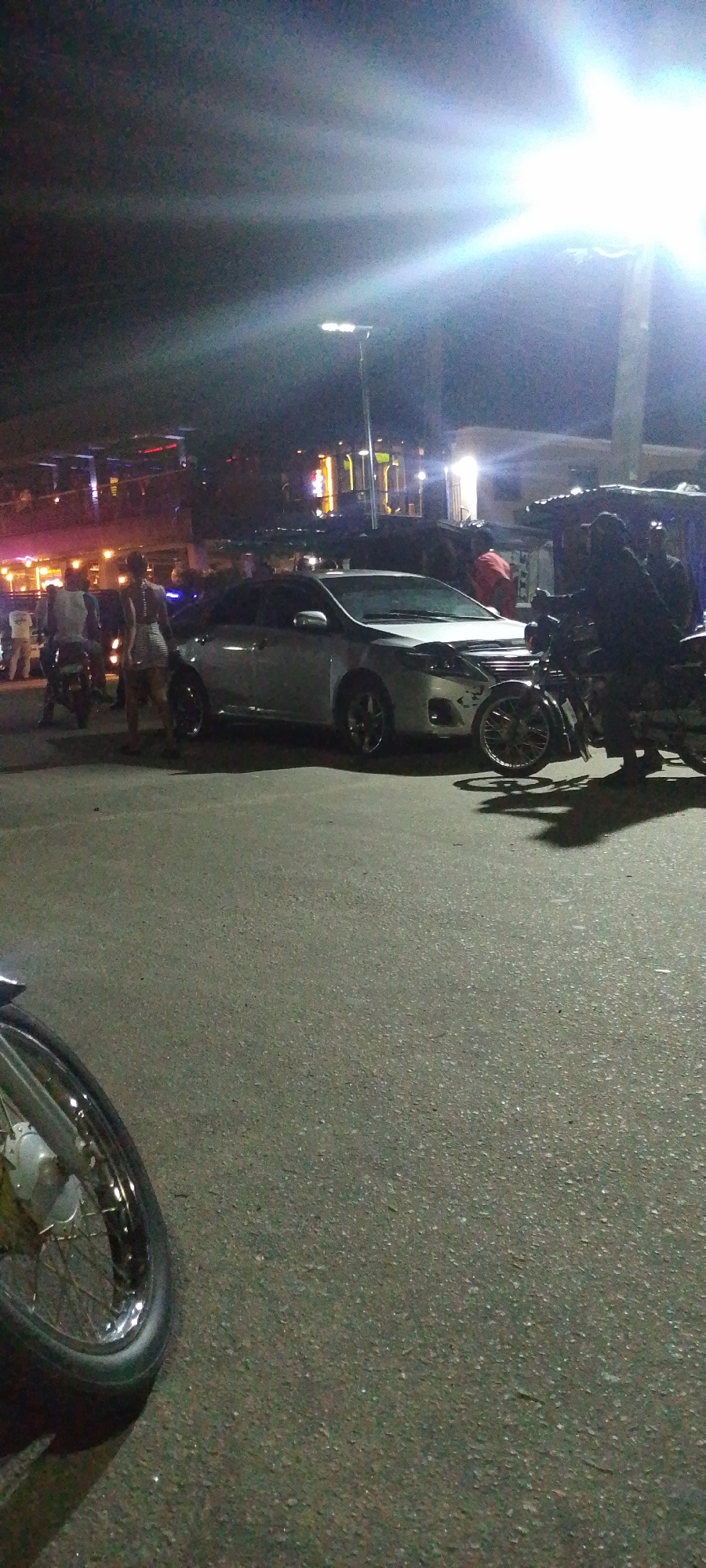 At night in Lokoja, Nigeria, a silver Toyota Corolla is parked on an asphalt road amidst pedestrian and motorcycle traffic. A woman in a light-colored dress stands near the car. Bright streetlights illuminate the scene. Buildings with lit signage are visible in the background. A motorcycle is positioned next to the car. The road surface appears wet and uneven.
