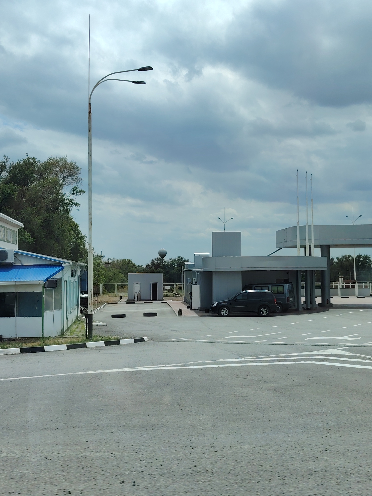 The image shows a checkpoint or entrance area. In the foreground is a paved road with white lane markings and a speed bump. To the left is a white building with a blue roof and windows. A tall pole with two streetlights stands near the building. In the middle ground, there is a grey building with a canopy where two vehicles are parked, a dark SUV and a van. There are also three flagpoles behind the canopy. Further back, there is a small grey building with two doorways and a water tower visible above the trees in the distance. The sky is overcast with clouds. The scene is set in an outdoor, possibly industrial or administrative area, located in the city of Eksperimental'nyi, Russia.