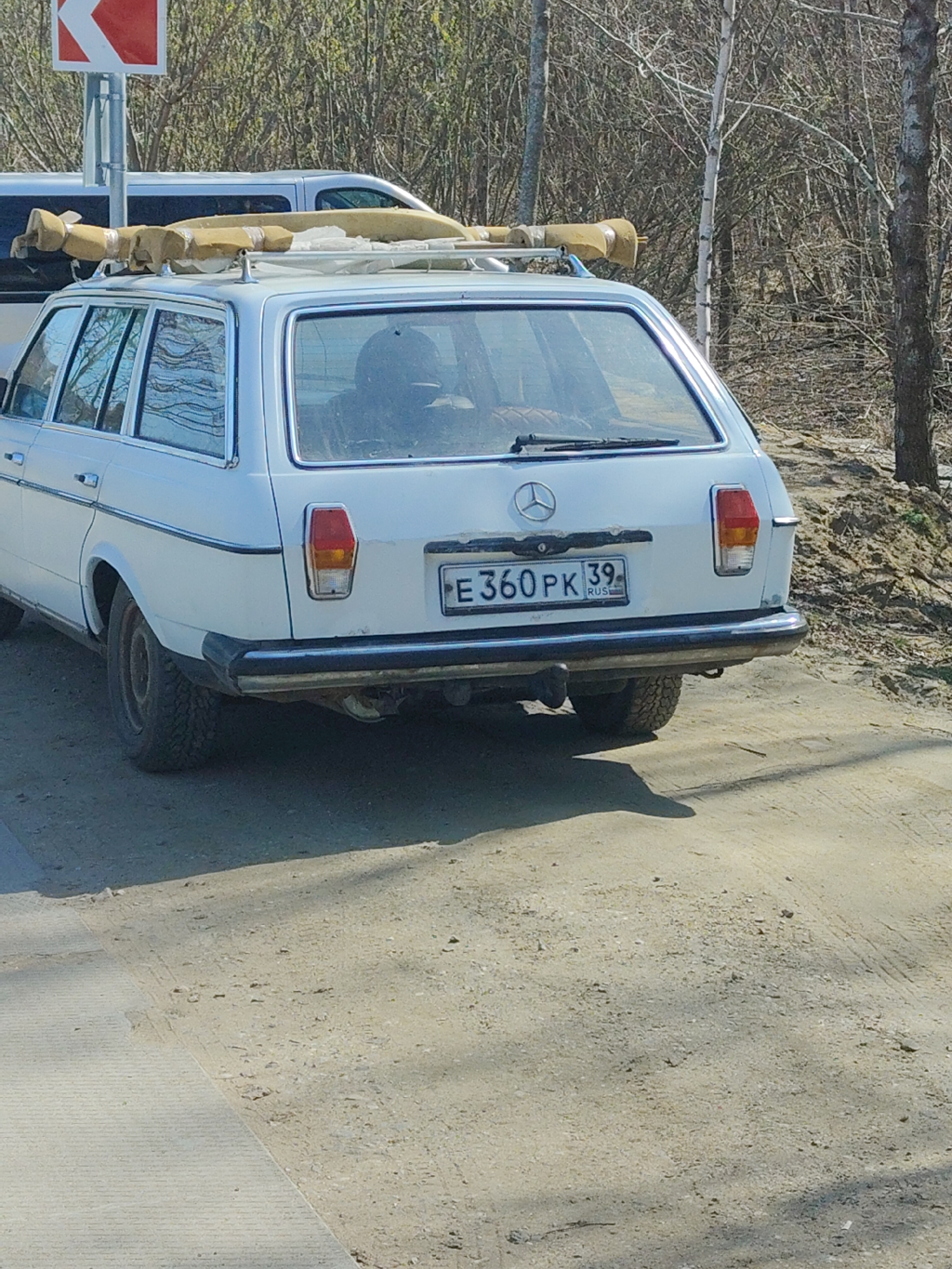 A white Mercedes-Benz station wagon with a roof rack containing wrapped and secured objects is parked on a dirt road next to a paved surface. The car has a Russian license plate that reads "E360PK 39 RUS". The background features a road sign, bare trees, and a grassy embankment. The location is likely in a rural or suburban area in Primorye, Russia, considering the presence of Russian license plates and the surrounding natural environment. There are no visible people directly interacting with the car.