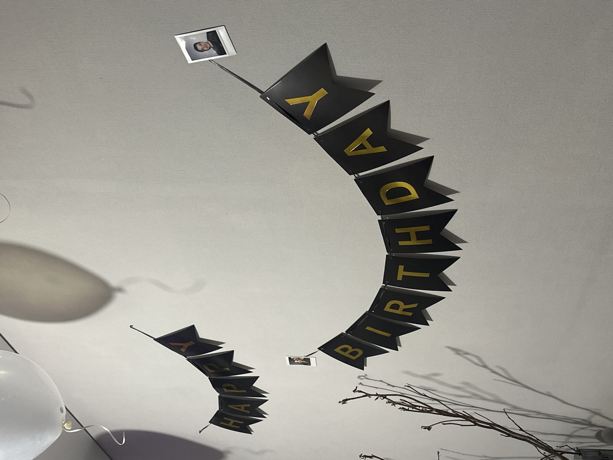 The image shows a "HAPPY BIRTHDAY" banner hanging from a white ceiling. The banner is made up of black pennants with gold letters. Small photographs are attached to the banner at two points. White balloons are visible in the lower left of the frame. Some decorative branches are in the lower right corner. The location is not discernible from the image.