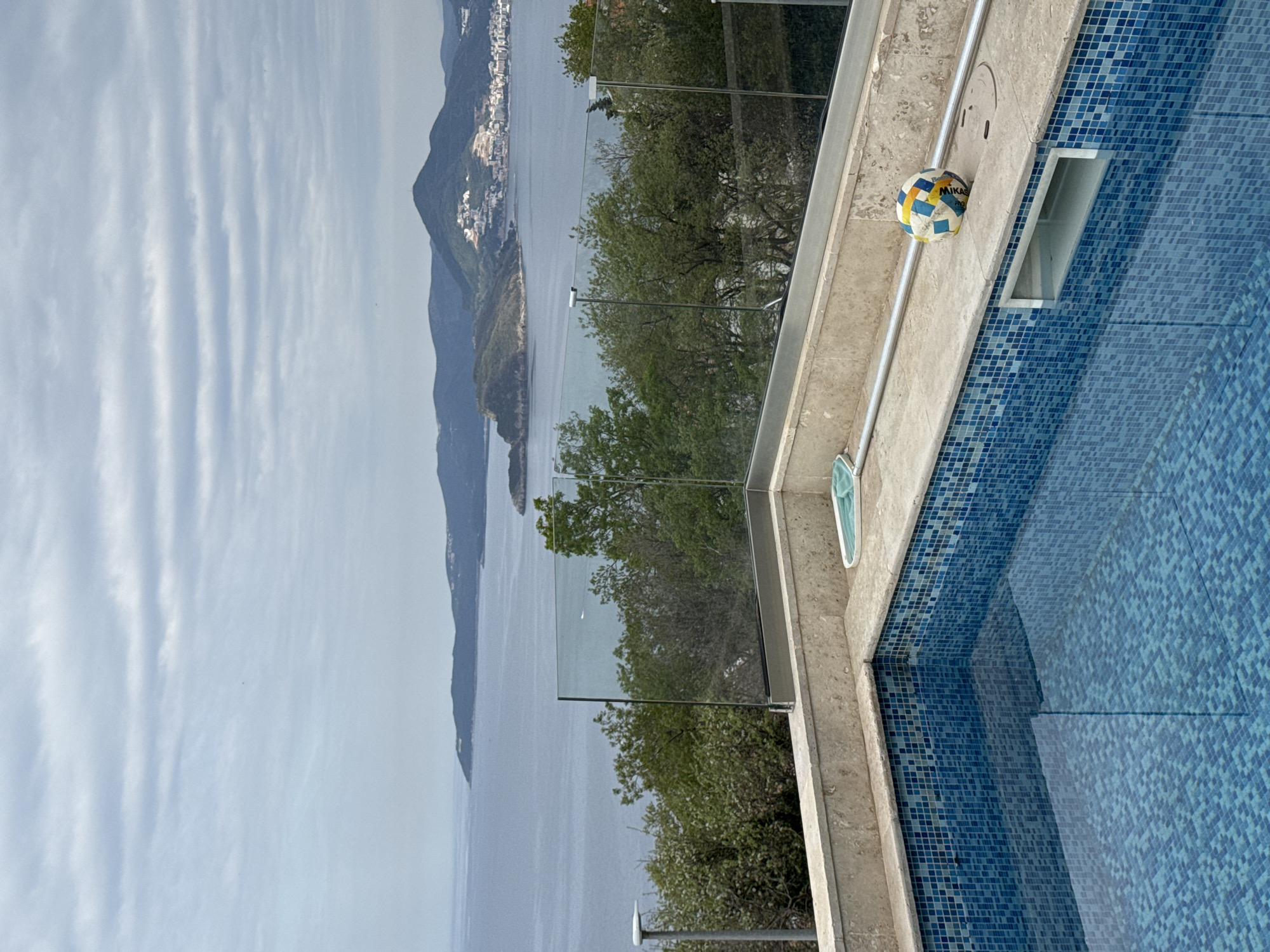 The image shows an outdoor scene likely taken in Blizikuće, Montenegro, featuring a swimming pool with blue mosaic tiles, a clear glass fence, and a view of the Adriatic Sea with mountains and coastline in the background. A Mikasa volleyball is present on the pool deck. The sky is partially cloudy. No people are visible.