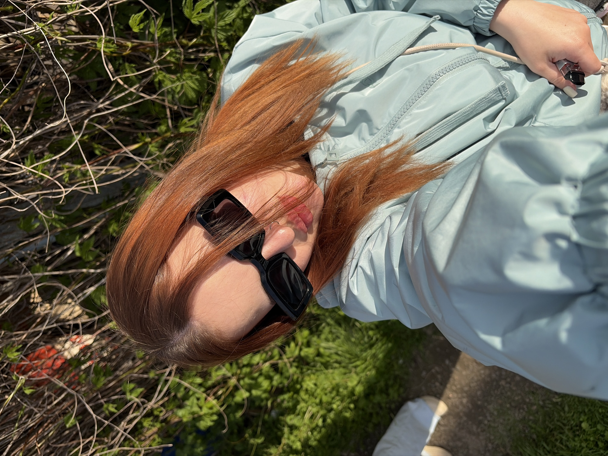 A woman with red hair and black sunglasses is lying in the grass near some twigs in Novopokrovskaya, Russia. She is wearing a light blue jacket and holding a small black object in her hand. A small portion of her shoe is visible at the bottom right of the image.
