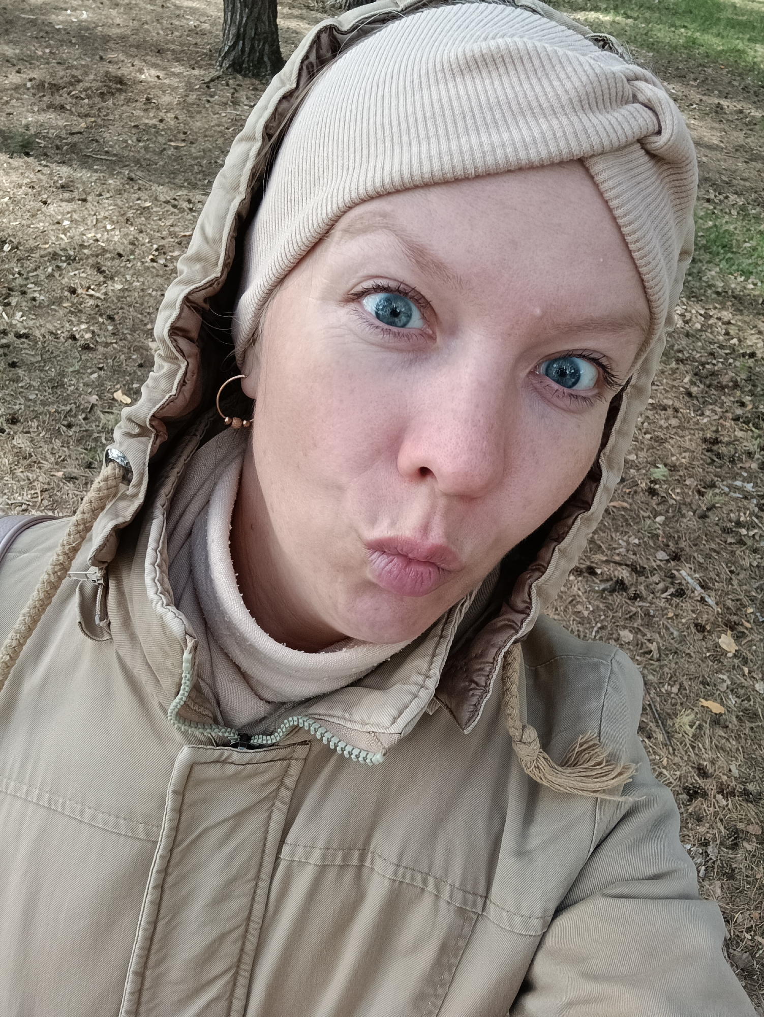 A fair-skinned woman with bright blue eyes is positioned in a close-up selfie shot outdoors. She is wearing a light tan or beige hooded jacket and a matching knit cap that covers her hair. She has a gold hoop earring in her left ear. The woman is making a kissing face, puckering her lips towards the camera. The background appears to be a wooded area with dry leaves and pine needles covering the ground. Bare tree trunks are visible behind her. The location could be a park or wooded area within the city of Chaikovsky, Russia.