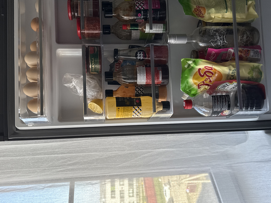 An open refrigerator is observed on the right side of the frame, adjacent to a window covered by sheer white curtains on the left. The window reveals an exterior view of multiple residential buildings under bright daylight.
Inside the refrigerator, the door shelves are visible. The top door shelf contains at least seven light brown eggs in a clear plastic egg holder. The second door shelf features a yellow half-lemon on the left, a clear plastic bag of unknown contents in the center, and several red-lidded jars, one with a "СКАЗКА" label, on the right. The third door shelf holds a tall yellow squeeze bottle with a black cap on the left, and various dark glass bottles, some labeled "ЧИМ ЧИМ" and "Tamaki," containing sauces or condiments. The lowest door shelf displays a clear plastic bottle with a red cap, partially filled with a dark liquid, next to a large green flexible pouch with Cyrillic text (likely "Ряба" mayonnaise or sauce). To its right stands a clear glass bottle with a frosted design, and a cream-colored flexible bag, possibly containing food.
No individuals or active interactions are present in the scene. The environment is an interior space, likely a kitchen or utility area, within a building overlooking a city or urbanized residential area.
