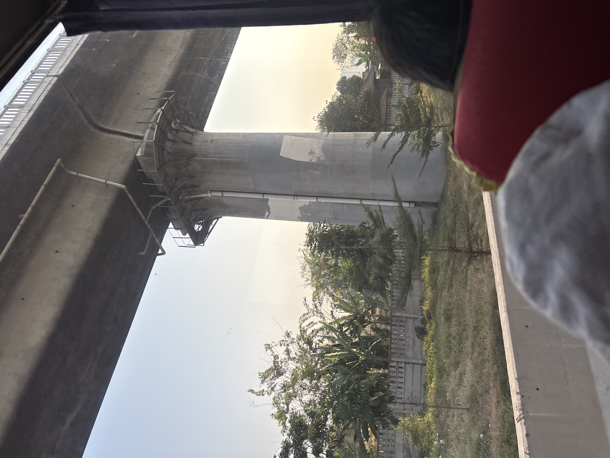 The image depicts an elevated concrete structure, likely a bridge or highway, casting a shadow over a rural landscape. A large concrete pillar supports the structure. Vegetation, including trees and grass, is visible below, with a concrete fence bordering the area. The sky is clear and pale, suggesting it might be early morning or late afternoon. In the foreground, parts of a vehicle's interior are visible, including the back of red seats and a person's head wearing a hat. The scene appears to be captured from inside a moving vehicle. The image captures a part of the infrastructure development in Jayamukti, Indonesia, showcasing the contrast between urban development and the natural environment.