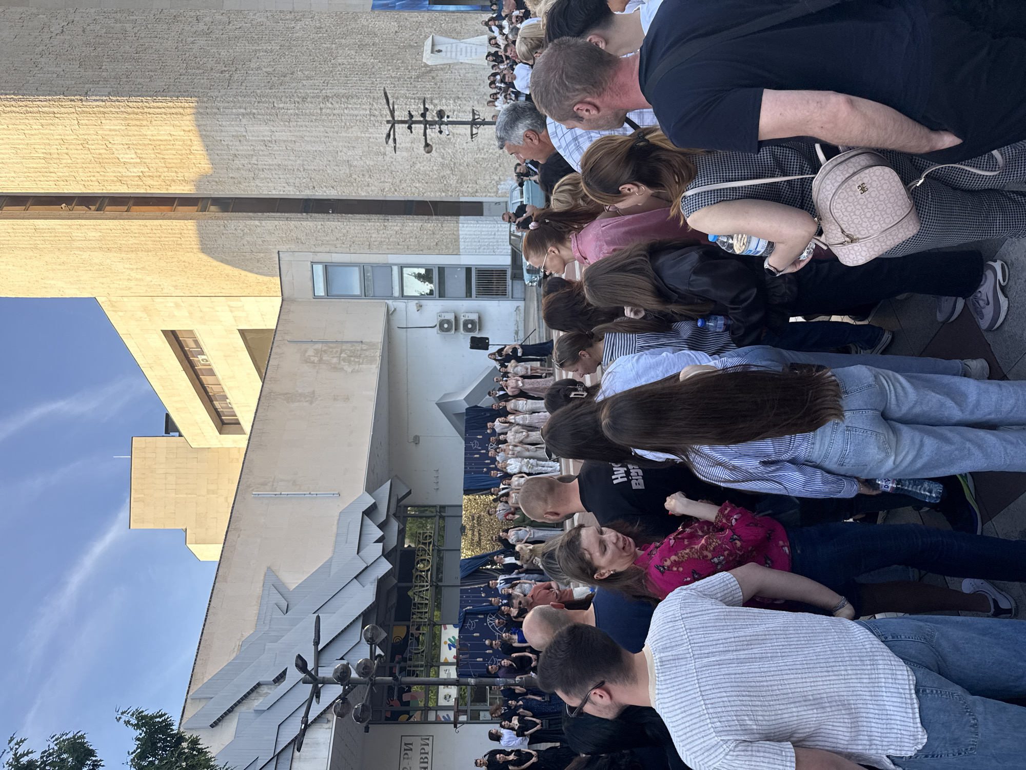 A crowd of people is gathered in front of the Blagoevgrad Drama Theater in Blagoevgrad, Bulgaria. The crowd is looking towards a stage where performers are dressed in matching outfits. The building behind the crowd is light-colored concrete with some decorative, angular elements. The sky above is blue with a few wispy clouds.