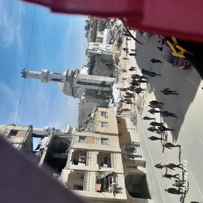 The image depicts a heavily damaged urban street scene in Khan Yunis, Palestinian Territory, under a clear blue sky with scattered clouds.
In the background, the Great Omari Mosque (Grand Mosque) stands prominently, characterized by its white/light-colored facade, dome, and a tall, cylindrical minaret. The mosque structure itself shows severe damage, with large sections collapsed and reduced to rubble around its base, though the minaret and dome remain partially intact but visibly damaged.
To the left of the mosque, a multi-story building exhibits extensive structural damage, particularly in its upper floors, with exposed rebar and concrete. On the lower, relatively intact balconies of this building, laundry is visible hanging. Debris piles are accumulated at the base of this structure and along the adjacent sidewalk.
The central and foreground areas feature a paved street bustling with numerous pedestrians, predominantly adults, with some children interspersed. They are walking in various directions, with a significant flow appearing to move towards the damaged mosque area. Individuals are dressed in civilian attire; some are observed carrying bags or bundles. A dark red sedan is partially visible parked on the right side of the street, obscured by a reddish foreground element. Utility lines stretch across the sky above the street.
The scene is viewed from an elevated perspective, partially framed by an indistinct reddish structure in the upper-left and right corners.