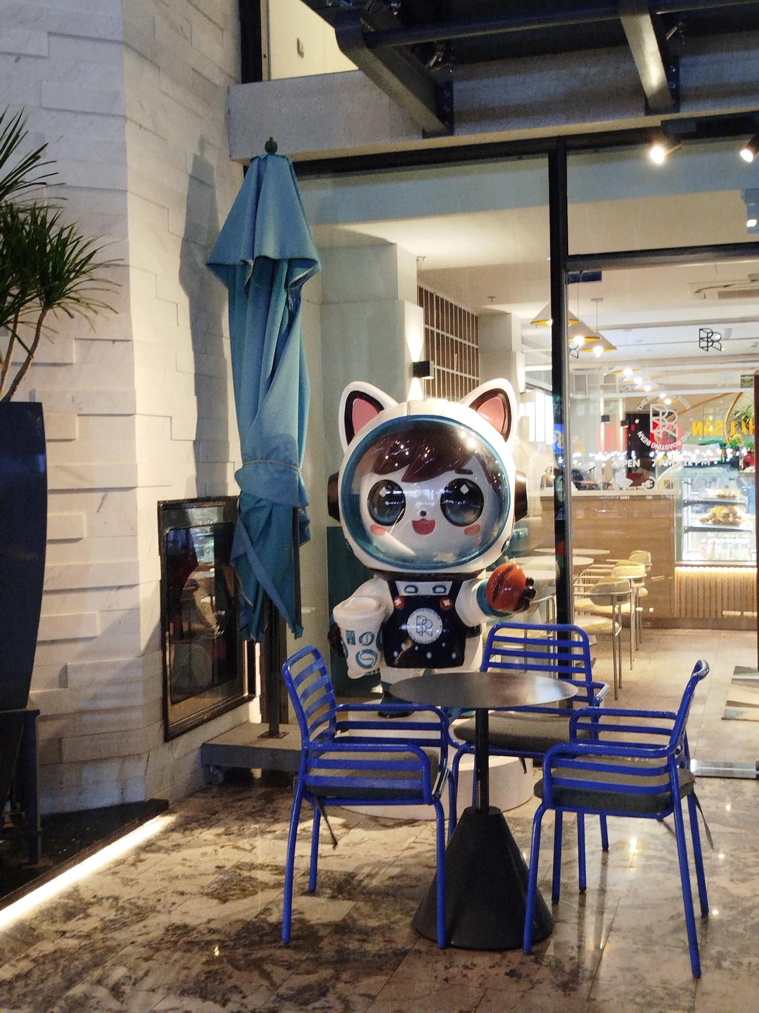 Outside a cafe in Ho Chi Minh City, Vietnam, a small, black round table surrounded by three blue chairs sits on a patterned, marble-like floor. Next to the table is a large, cartoonish statue of a cat-like figure in an astronaut suit holding a coffee cup. An unused blue umbrella is furled and leaning against a pillar to the left of the statue. A glass window behind the statue provides a view into the cafe's interior, showing tables, chairs, and a display case with food items. The cafe's signage is visible, though the text is partially obscured by reflections. To the left, a tall potted plant stands against a textured white wall, with a framed picture or advertisement mounted nearby. A strip of light is embedded in the floor, casting a bright glow.