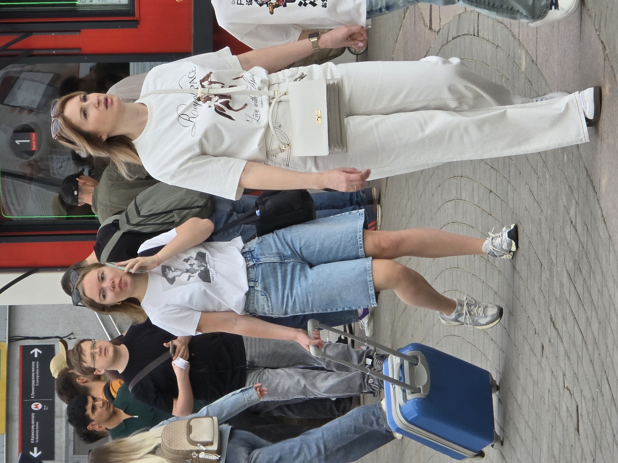 A medium shot captures a crowd of people near the entrance of a train in Moscow, Russia. A young woman in the center is on her phone while pulling a blue suitcase. She wears a white t-shirt with a portrait, denim shorts, and sneakers. Behind her is a man in a black t-shirt. To the right and above her is another woman dressed in white jeans and a t-shirt with a bag over her shoulder. The people are standing on a paved surface. A red train is visible in the background. Signage in Cyrillic script and English can be seen near the woman.