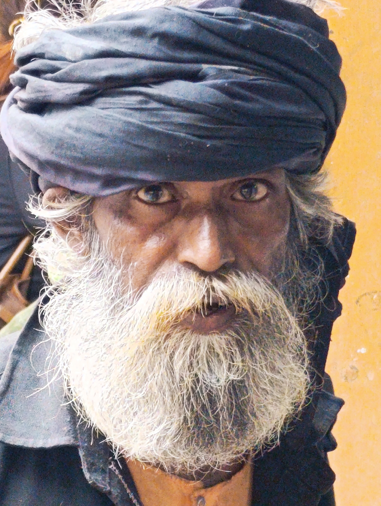 The image shows a close-up portrait of an elderly man with a long, white beard and hair peeking out from under a dark blue turban. He is wearing a dark jacket or shirt. The man has deep-set eyes and wrinkles on his face, suggesting an aged appearance. The background is a plain, textured yellow wall. The lighting is slightly harsh, casting shadows and highlighting the man's features. There is no discernible indication of the location being Manali, India, or any specific urban setting. The image appears to be a candid or posed portrait of an individual.