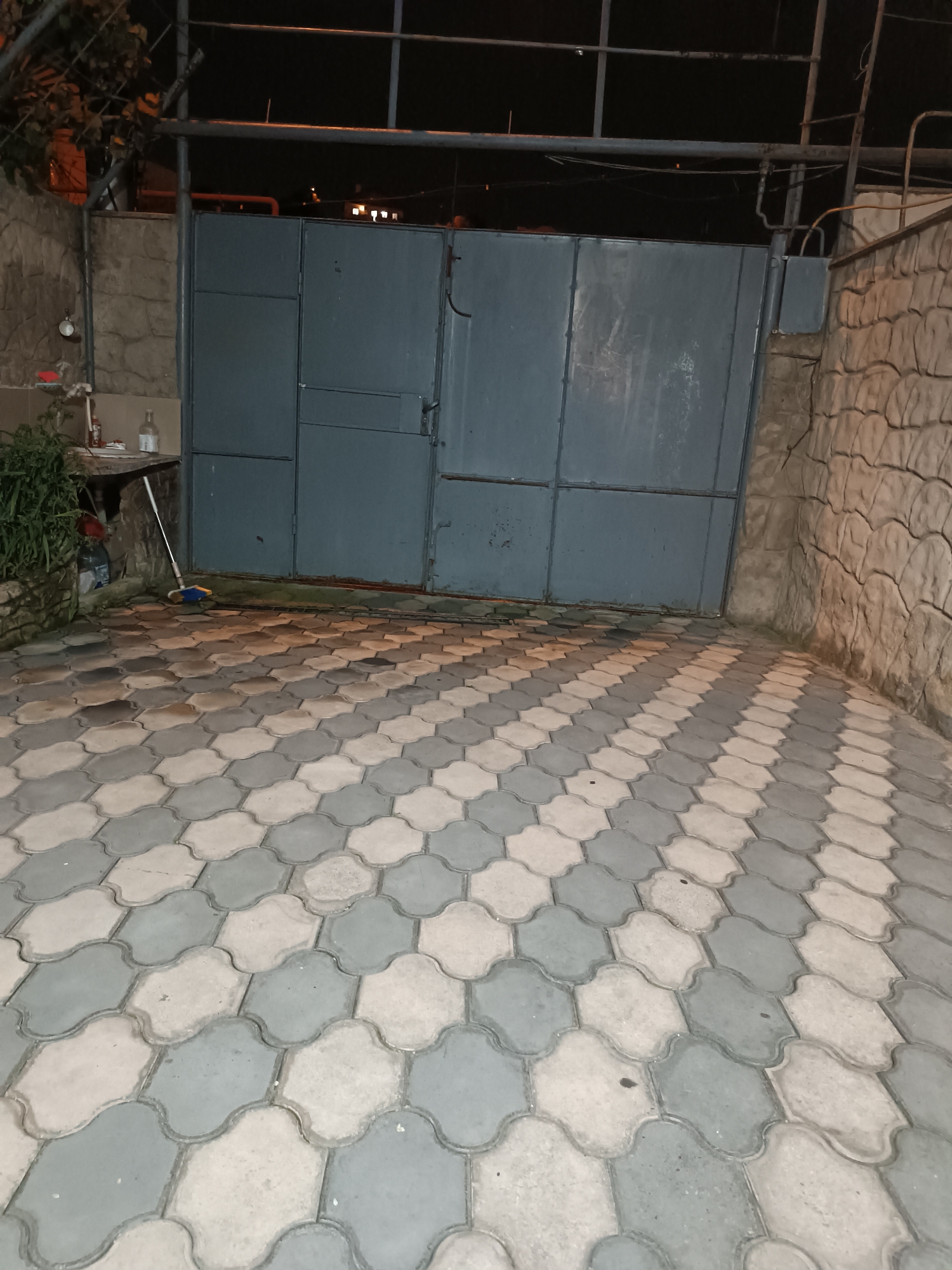 A blue metal gate is centrally located in the image. To the left of the gate, a stone wall is visible with some greenery and a sink area. To the right of the gate, another stone wall with a textured pattern is present. The ground in front of the gate is paved with interlocking hexagonal stones in a grey and beige pattern. The scene appears to be an outdoor entrance or driveway, possibly at night, given the lighting and dark background. The overall impression is of an exterior utilitarian space. The specific location in Tbilisi, Georgia cannot be determined from the image.
