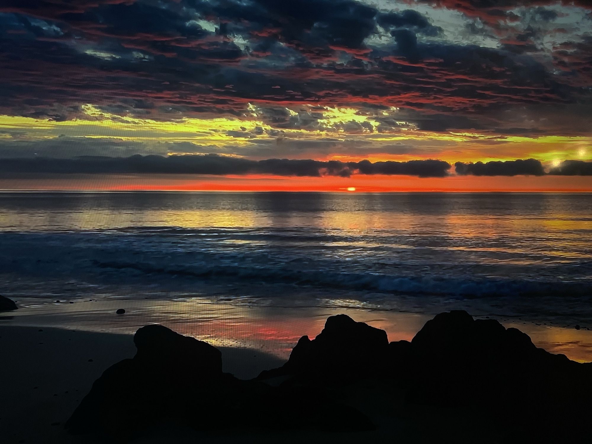 This image depicts a breathtaking sunrise or sunset over a vast ocean, characterized by a dramatic and colorful sky. The horizon line bisects the image, with the sun, a bright orange disc, peeking just above it and slightly left of center. A low band of dark clouds partially obscures the sun.
Above the horizon, the sky is filled with layered, textured clouds that transition from dark grays and blues in the upper regions to fiery yellows, oranges, and deep reds and pinks closer to the sun. The lower clouds near the horizon also catch the warm light, glowing with intense hues.
The ocean surface below the horizon reflects the vibrant colors of the sky, particularly the yellows, oranges, and reds, creating shimmering pathways on the water. Gentle waves are visible breaking closer to the shore, with the wet sand of the beach also mirroring the colorful light. In the immediate foreground, large, dark rock formations are silhouetted, framing the bottom of the scene and creating a strong contrast against the bright sky and reflective water. Smaller pebbles are scattered on the wet sand.
The scene is tranquil and devoid of human activity, focusing entirely on the natural beauty of the coastal landscape at either dawn or dusk. No text is visible in the image.