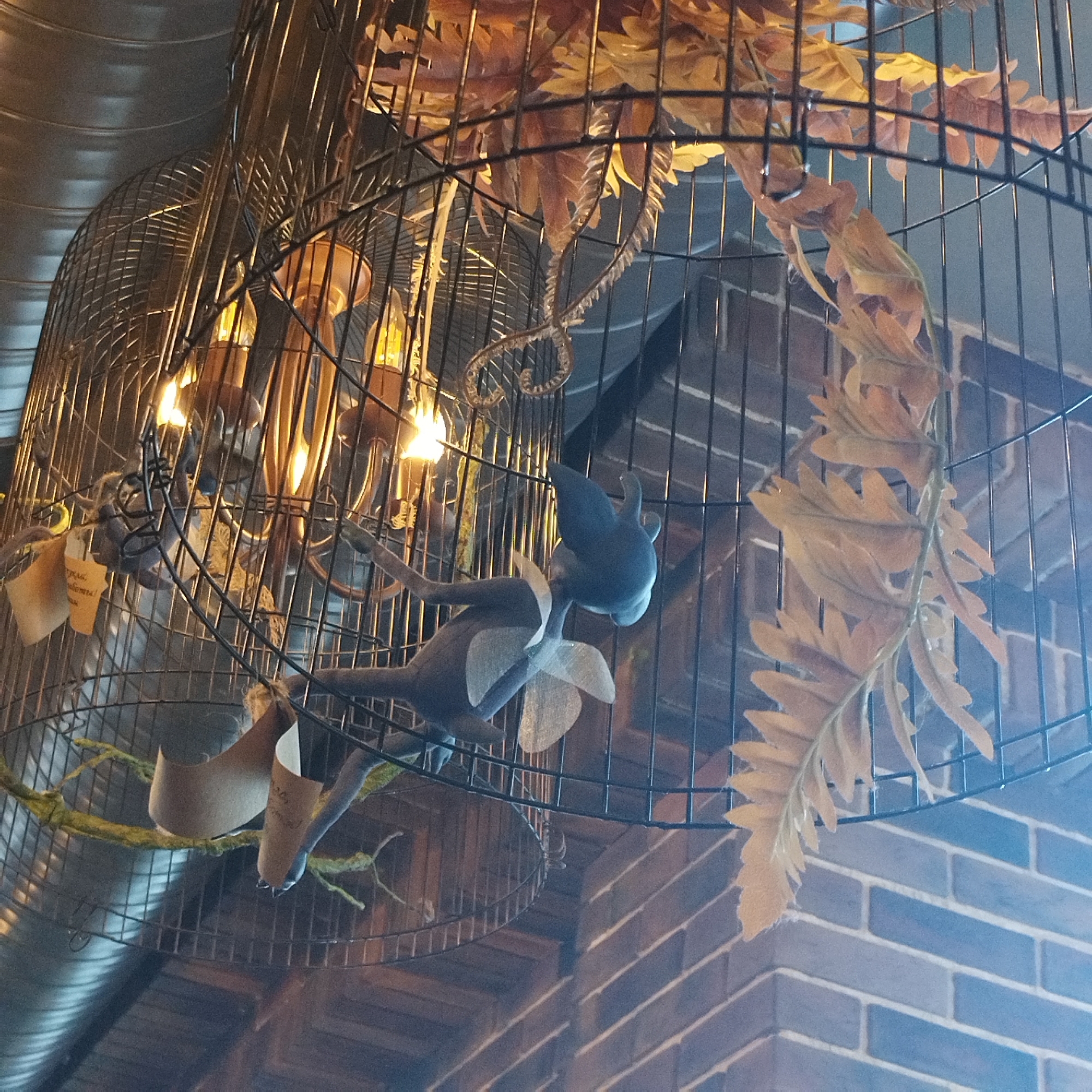 An indoor scene showcases an elaborate decorative installation featuring multiple large, black metal birdcages, possibly repurposed as light fixtures, hanging from a ceiling. Inside the cages, artificial autumn foliage in shades of orange and brown is intertwined with branches resembling mossy green twigs. Several bare light bulbs emit a warm, orange-yellow glow, illuminating the cage's interior.
A whimsical, blue-grey plush figure, resembling a small winged creature with large ears and a light-colored bow around its neck, hangs suspended within one of the cages. Another element is a delicate, coiled decorative piece, possibly a stylized vine or tail, made of a lighter, reflective material. From a mossy branch, a rolled-up piece of light brown paper, possibly a scroll, hangs with illegible handwritten text.
The background reveals a textured environment. To the upper left, a silver-colored, corrugated metal air duct runs horizontally. Below and to the right, a brick wall is visible, with dark reddish-brown bricks on the left side and transitioning to a blue-grey brick pattern on the right, suggesting varying wall sections or lighting effects. There is no discernible activity or event taking place; the image captures a static decorative display, likely in a cafe, restaurant, or similar establishment, emphasizing an eclectic and cozy ambiance. The artificial lighting suggests it could be evening or a dimly lit interior.
