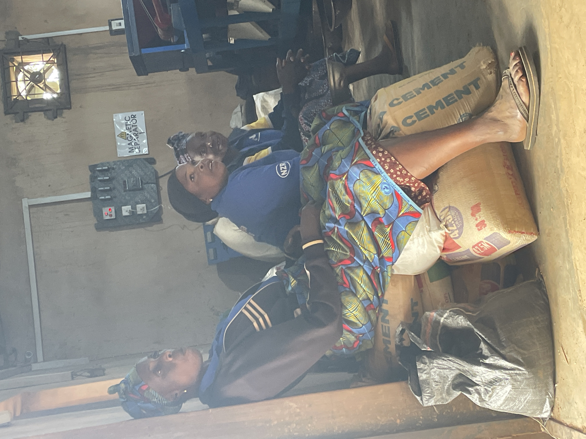 In Gura Top, Nigeria, three women are seated on bags of cement inside a dimly lit, dusty building. The woman on the left, wearing a blue jacket and a headscarf, looks to her right. The woman in the center wears a blue shirt with an "NZR" logo and a patterned skirt, and she is looking forward. The woman on the right, partially obscured, is also looking forward. In the background, industrial equipment, including a control panel labeled "MAGNETIC SEPARATOR," and a fan mounted on the wall are visible. Bags of cement and a black plastic bag are piled on the floor.