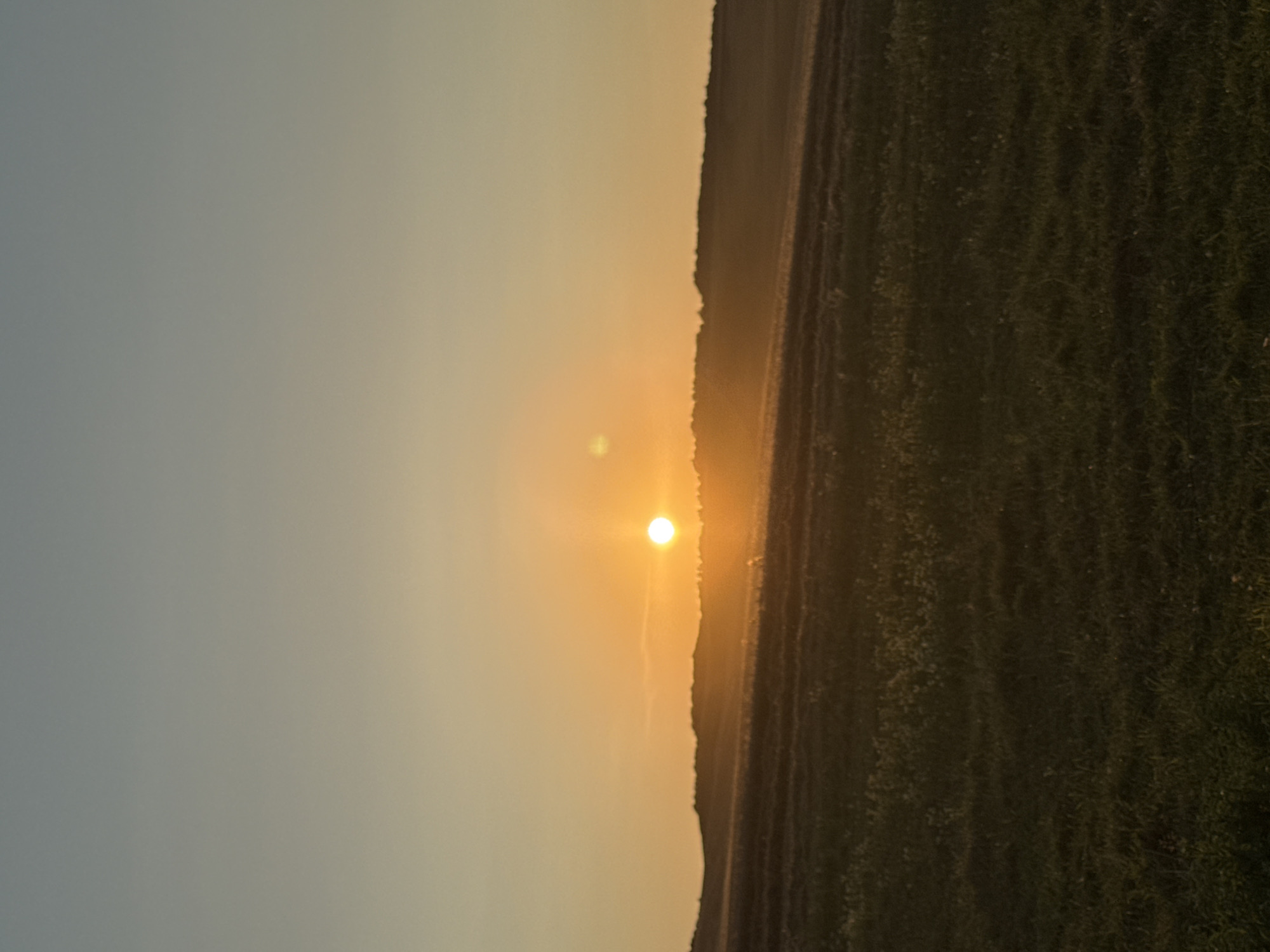A wide, horizontal shot captures a tranquil sunset over a rural landscape. The sun, a bright, glowing orb, is positioned in the upper-middle of the frame, casting a warm, orange light across the scene. The sky transitions from a pale blue at the top to a soft yellow and orange hue near the horizon, indicating the setting sun. Below the sky, a dark, undulating silhouette of hills or a treeline forms the horizon. In the foreground, a vast, grassy field is visible, with its texture and subtle variations in color suggesting gentle slopes and patches of shorter and longer grass. The lighting creates a soft, diffused effect, with minimal sharp details, contributing to the peaceful atmosphere of the image. The location is identified as Unknown, Unknown.