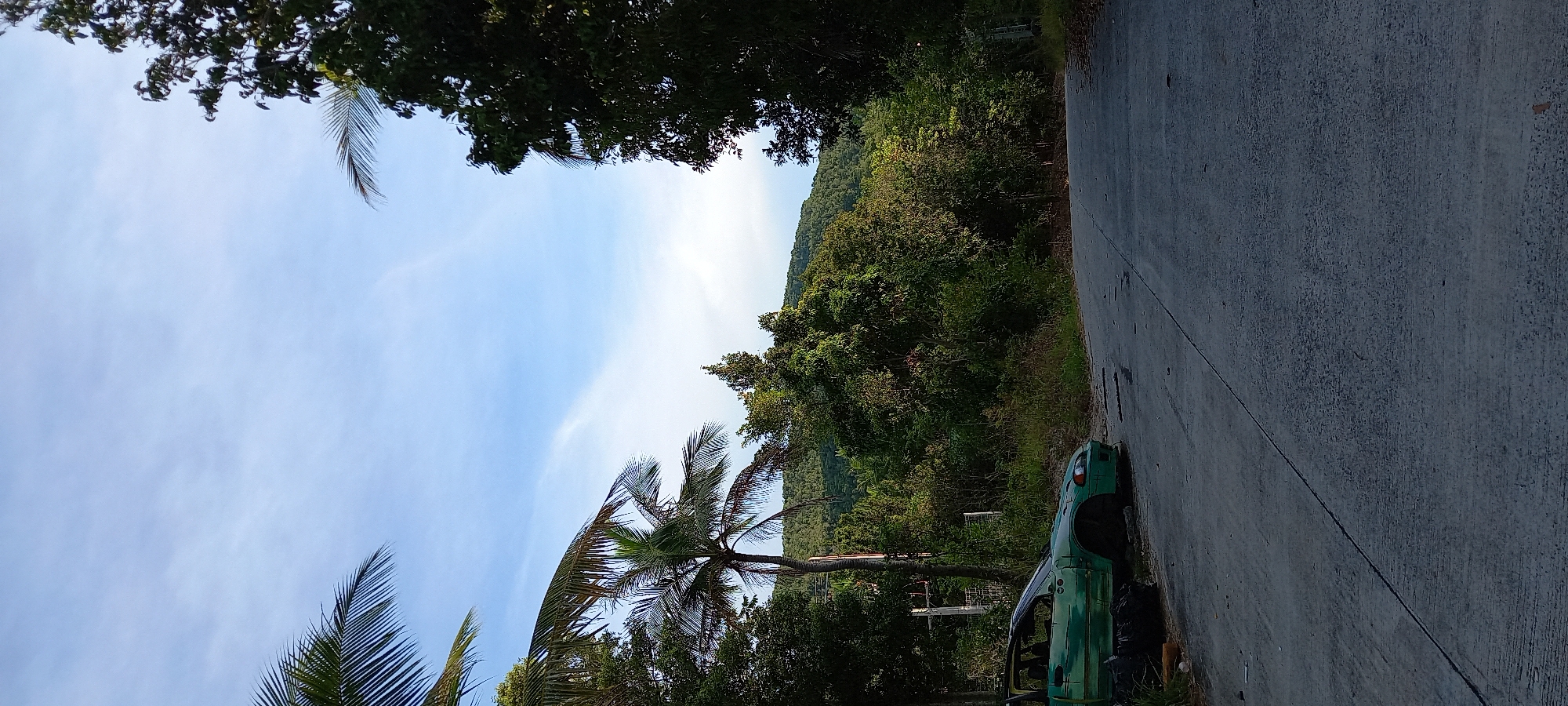 The image shows a roadside scene in Baan Wang Ta Kien, Thailand. The sky is light blue with scattered clouds. Trees and vegetation cover the background, including a small mountain visible in the distance. In the foreground, a weathered concrete road runs vertically. Adjacent to the road on the right, a heavily damaged and decaying green vehicle rests amidst the surrounding foliage. The vehicle appears to be missing doors and windows, showing significant signs of disrepair. Palm trees are also present along the left side of the road. The image is taken from a low angle.