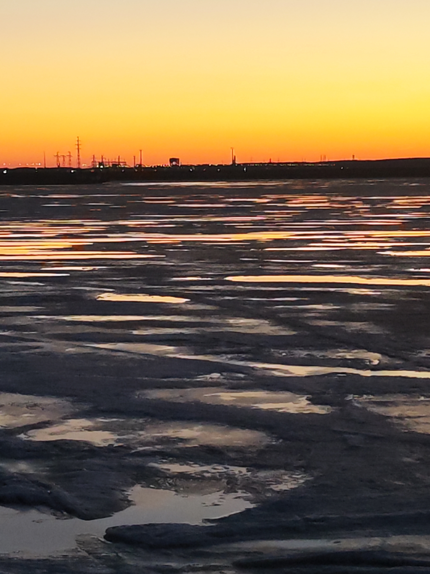 The image depicts a landscape at dusk or dawn, likely a tidal flat or shallow water area. The foreground is dominated by mudflats or sand with scattered water puddles or channels reflecting the light from the sky. The water is relatively still, creating elongated reflections.
In the mid-ground, the water extends horizontally, with the same reflective qualities. A distant horizon is visible, demarcated by a dark, uneven line, possibly a coastline or distant buildings. Silhouetted structures, possibly industrial or urban elements such as power lines, factories, or buildings, are faintly visible against the sky.
The sky occupies the upper portion of the frame, featuring a gradient of colors, ranging from a bright yellow or orange near the horizon to a dimmer shade further up. The overall lighting suggests either sunrise or sunset, creating a warm, soft glow.
The specific location within the city of Unknown is undetermined due to lack of definitive features. There are no people visible in the image.