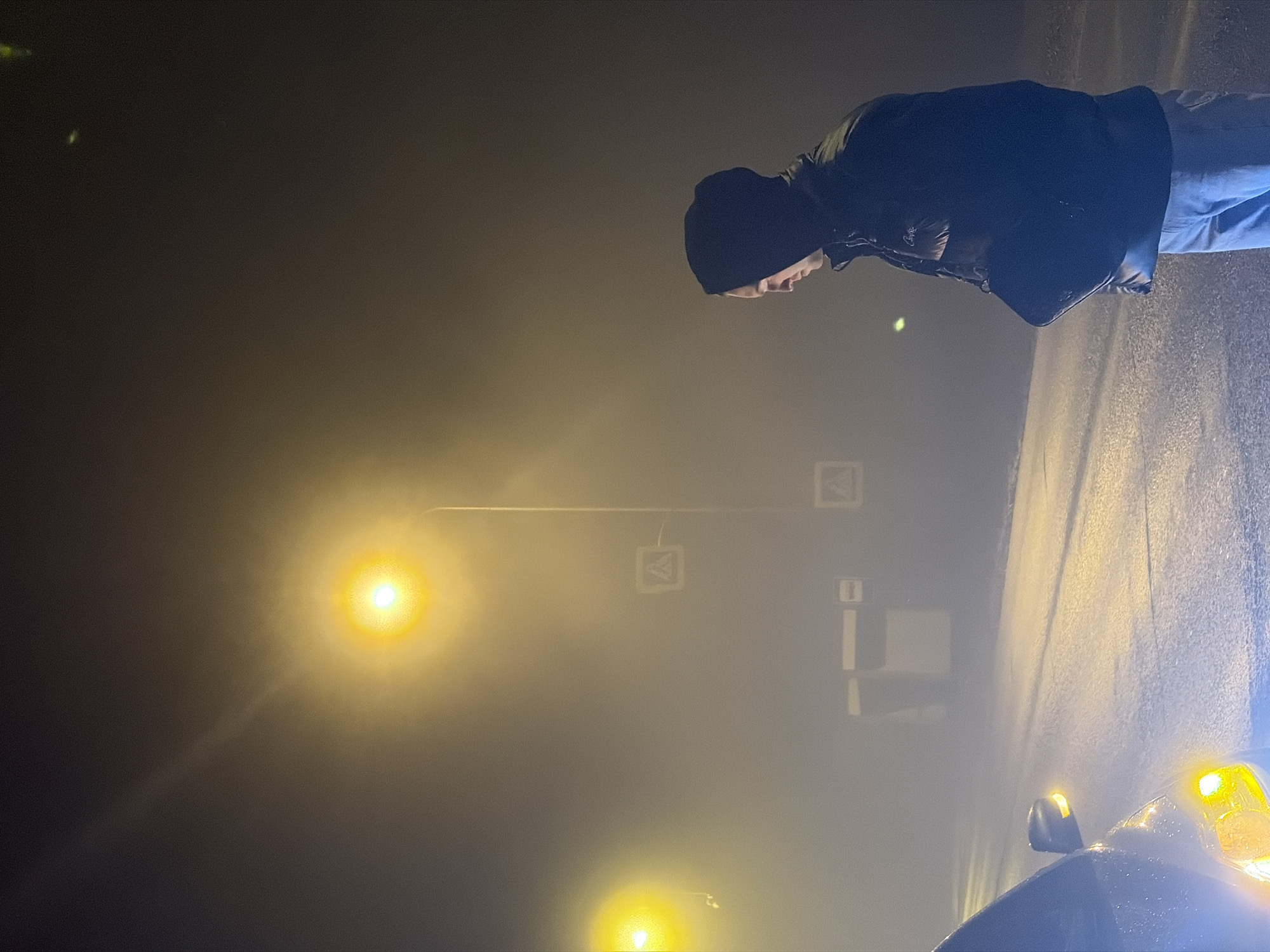 A man wearing a black beanie, a black jacket, and blue jeans stands on a road in heavy fog at night. A car's headlights illuminate the fog in the lower right of the frame. Streetlights glow in the background. Traffic signs are visible on the street. The scene appears to be a street in Noviy, Russia, judging by the signs, the streetlights, and the fog typical of the region. The man is facing forward and appears to be looking down.