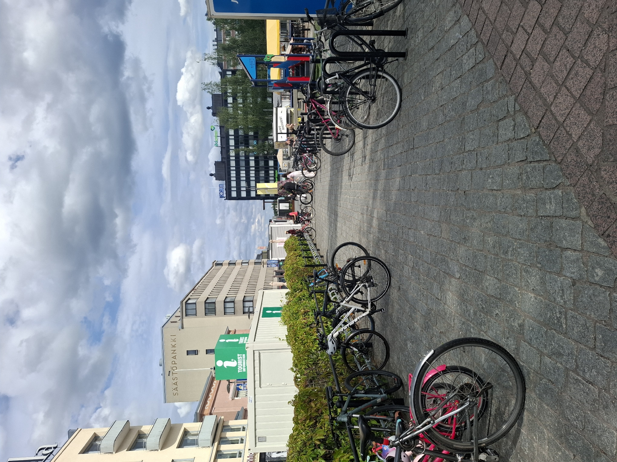 In Mikkeli, Finland, this image captures a sunny day with a cloudy sky. The scene is an urban plaza with cobblestone paving. On the left, a light yellow building with balconies is visible, followed by a pink building and a white container labeled "TOURIST INFORMATION" with green signage. A row of bicycles is parked in front of the container, and more bicycles are lined up further into the plaza. Several people are seen interacting with the bicycles, and some are walking in the background. The plaza also features a playground structure with yellow and blue elements. The overall atmosphere is that of a public space with recreational and informational facilities.