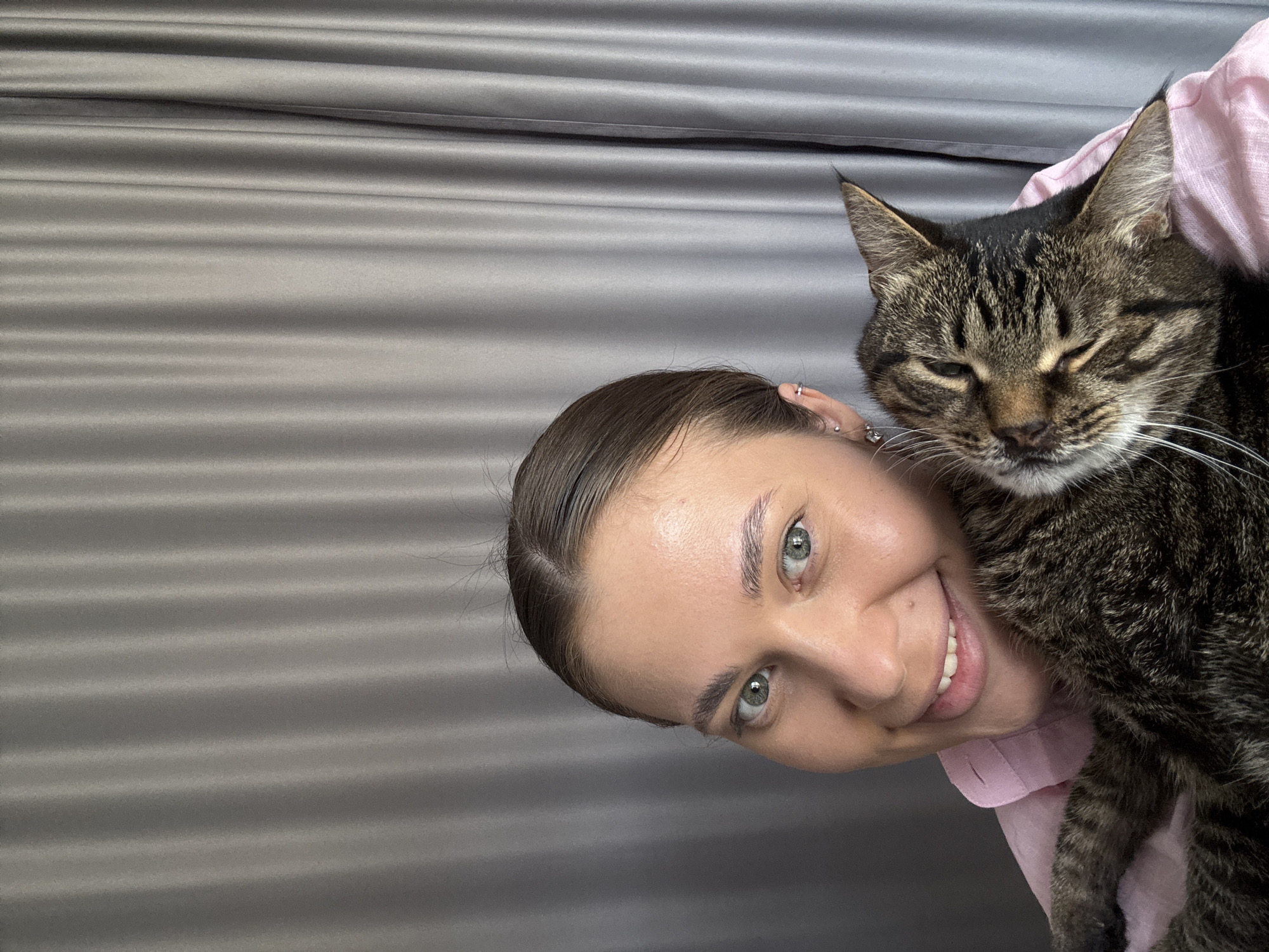 The image features a young woman smiling and holding a tabby cat close to her face, suggesting a warm and affectionate moment. The woman's head is tilted, giving the impression of a selfie or an informally captured shot.
The woman has light-colored eyes (likely green or hazel), with visible eyebrows and a broad, open-mouthed smile showing her teeth. Her hair, which appears dark brown, is pulled back, and she wears multiple small, silver-toned earrings on her visible ear. She is wearing a light pink garment, possibly a shirt. Her expression conveys happiness and contentment.
The cat is a brown tabby with distinct dark stripes and markings. It is resting comfortably, with its eyes mostly closed, suggesting a state of relaxation or drowsiness. Its white whiskers are prominent. The cat's face is positioned very close to the woman's, almost touching her cheek.
The background consists of a textured surface with horizontal, undulating ridges, appearing in a muted grey color. This backdrop is uniform and simple, putting the focus entirely on the woman and her cat. The lighting is soft and even, indicating an indoor setting without harsh shadows. There is no visible text in the image.