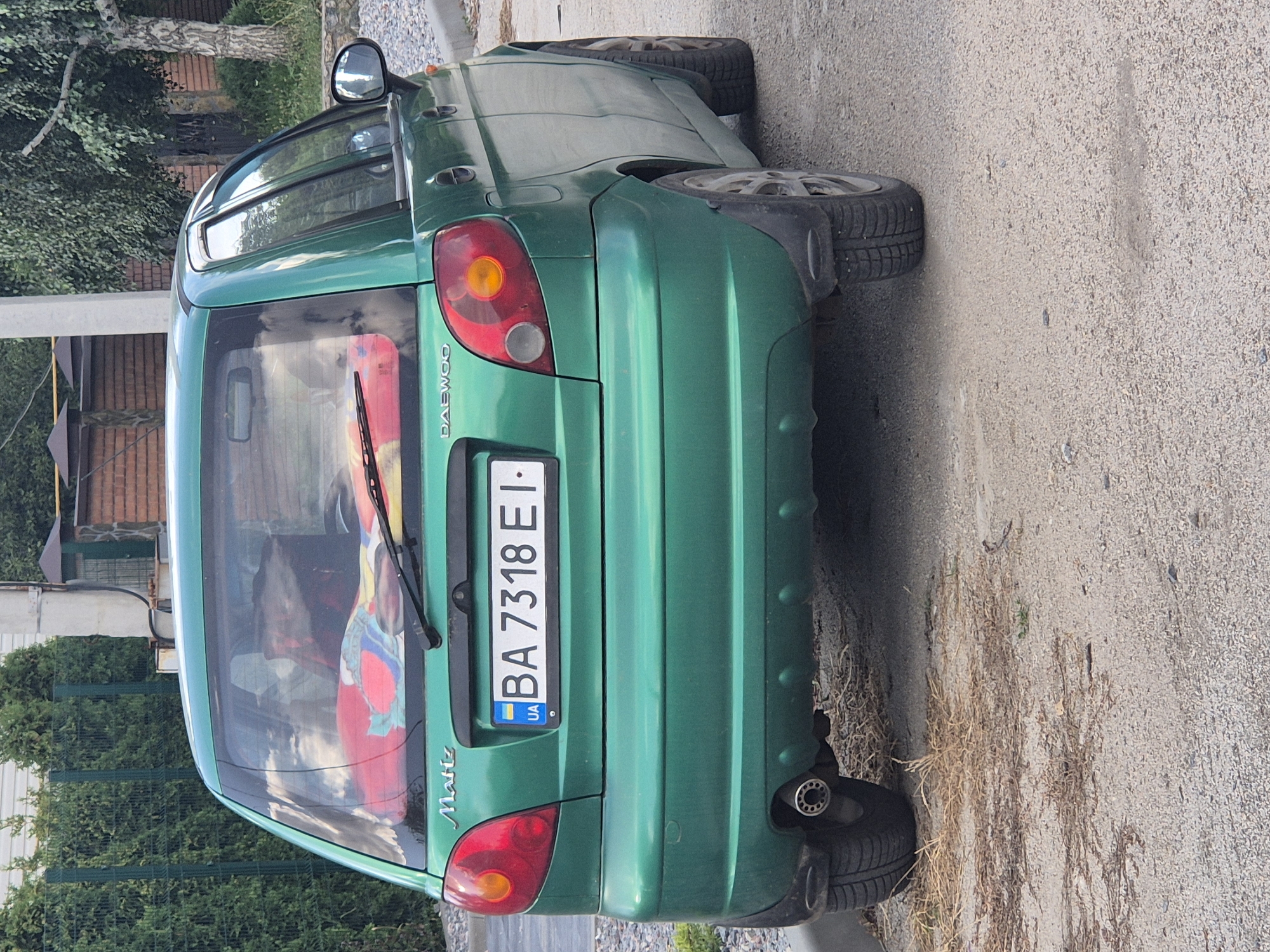 A rear, eye-level, medium shot captures a green Daewoo Matiz hatchback parked on a gravel surface in Sozonivka, Ukraine. The car is oriented towards the viewer, facing away from the camera. The vehicle's license plate, "BA 7318 EI," is clearly visible. The car's interior shows a red and colorful patterned blanket draped over the rear seats. The background features a green hedge, a brick fence, and trees. The lighting suggests it is daytime, with the sun casting slight shadows. There are no people visible in the scene, and the car appears to be stationary.