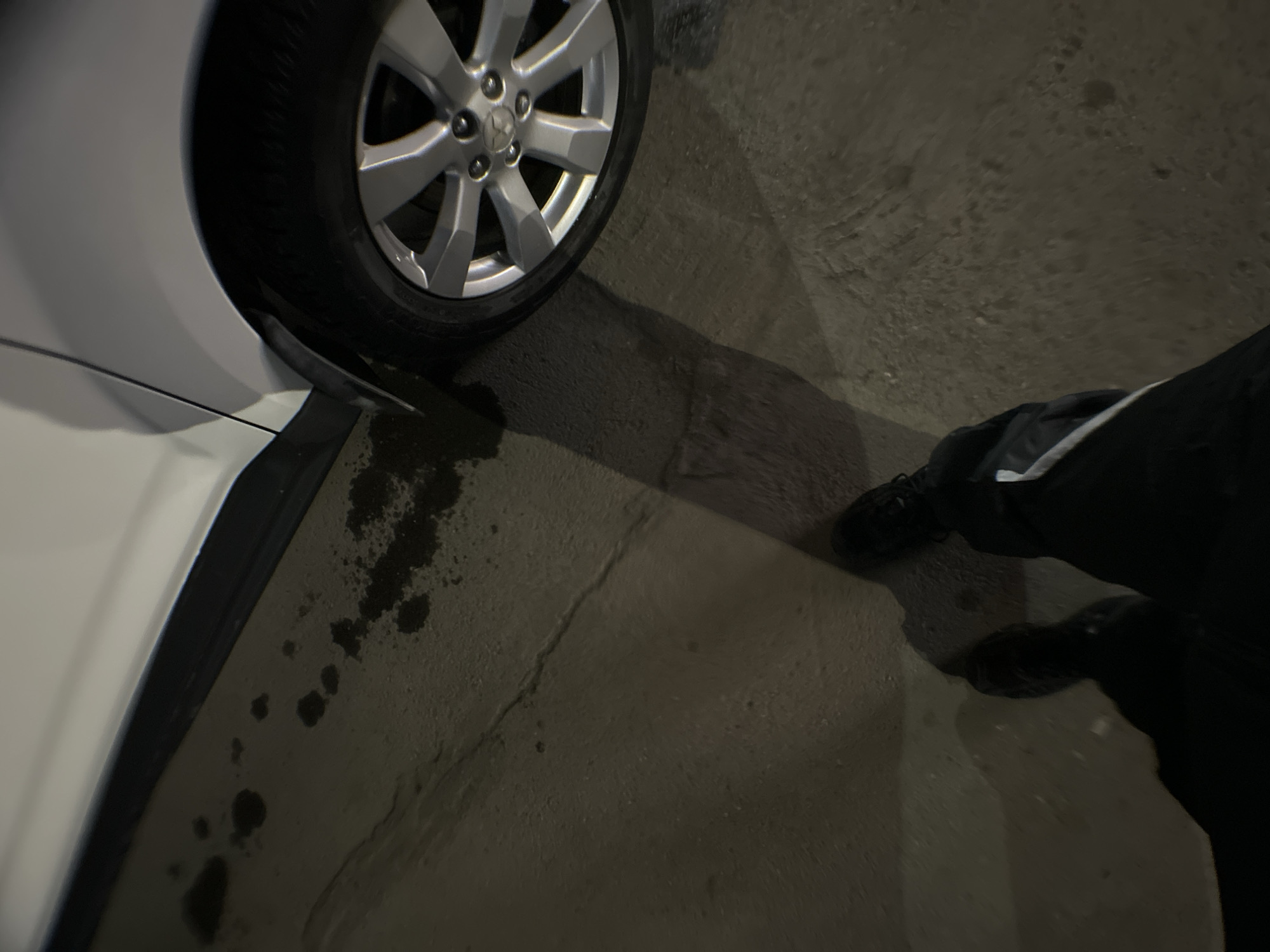 The image depicts a partial view of a white car, focusing on the front driver's side tire and a portion of the car body. The tire is black with a silver rim. The ground is concrete and stained with dark spots resembling oil or water. A person's legs are visible on the right side of the frame. The person is wearing dark pants with a light-colored stripe and dark shoes. The photograph appears to have been taken at night or in a poorly lit environment. The location, Chaikovsky, Russia, is unknown as there is no information in the image to suggest it.
