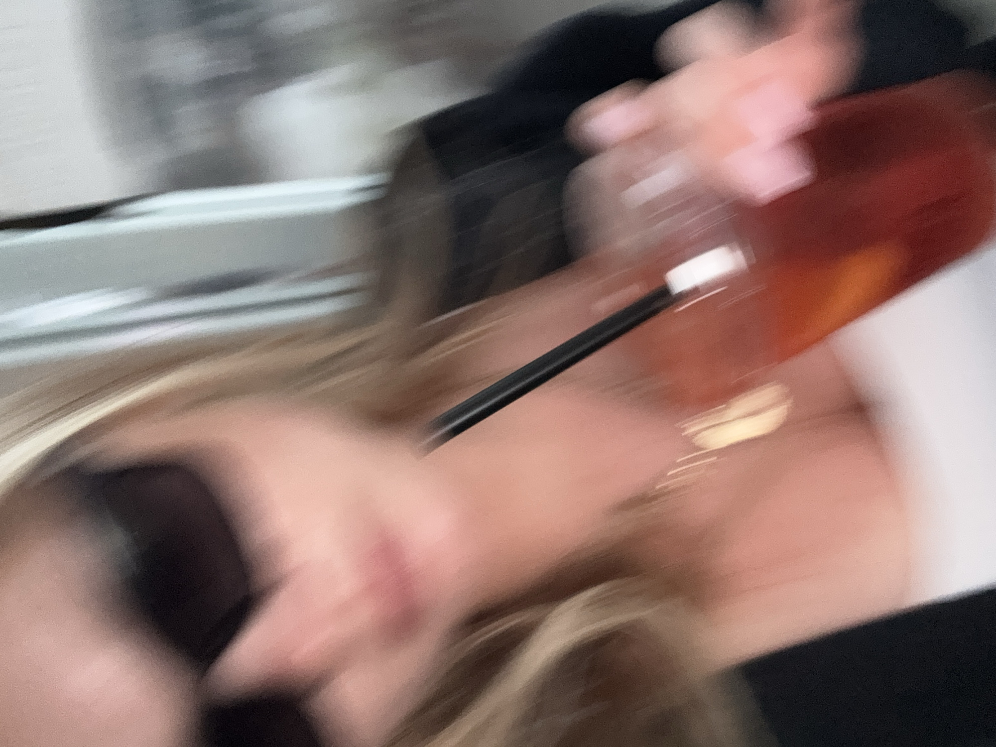 A blurry image shows a person holding a drink with a straw. The drink appears to be amber-colored. The person has blonde hair and is wearing a dark-colored top. The background is out of focus, but a window or bright surface is visible on the left. There is no information to indicate the location is Tel-Aviv, Israel.