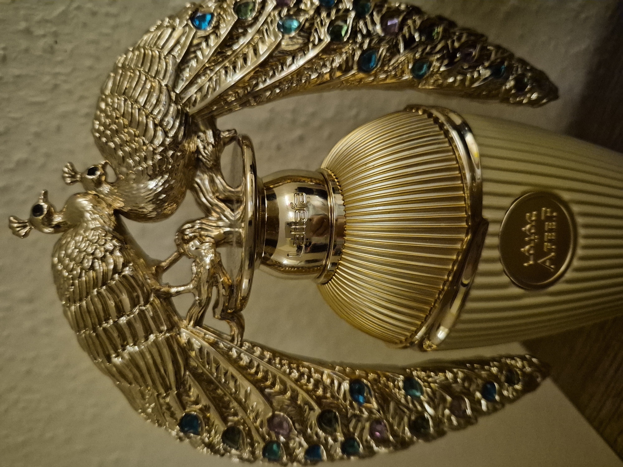 A close-up, eye-level shot shows the top of a gold-colored perfume bottle with elaborate peacock-shaped embellishments. Two gold peacocks are perched on a branch atop the bottle's cap, their iridescent feathers extending outwards and adorned with small blue, green, and purple gemstones. The bottle itself is cylindrical and ridged, with gold lettering visible on the neck and a circular emblem on the body. The background is a neutral, textured wall. The lighting is warm and soft, highlighting the metallic sheen and jewel tones of the perfume bottle. The image is focused on the ornate details of the bottle's design.