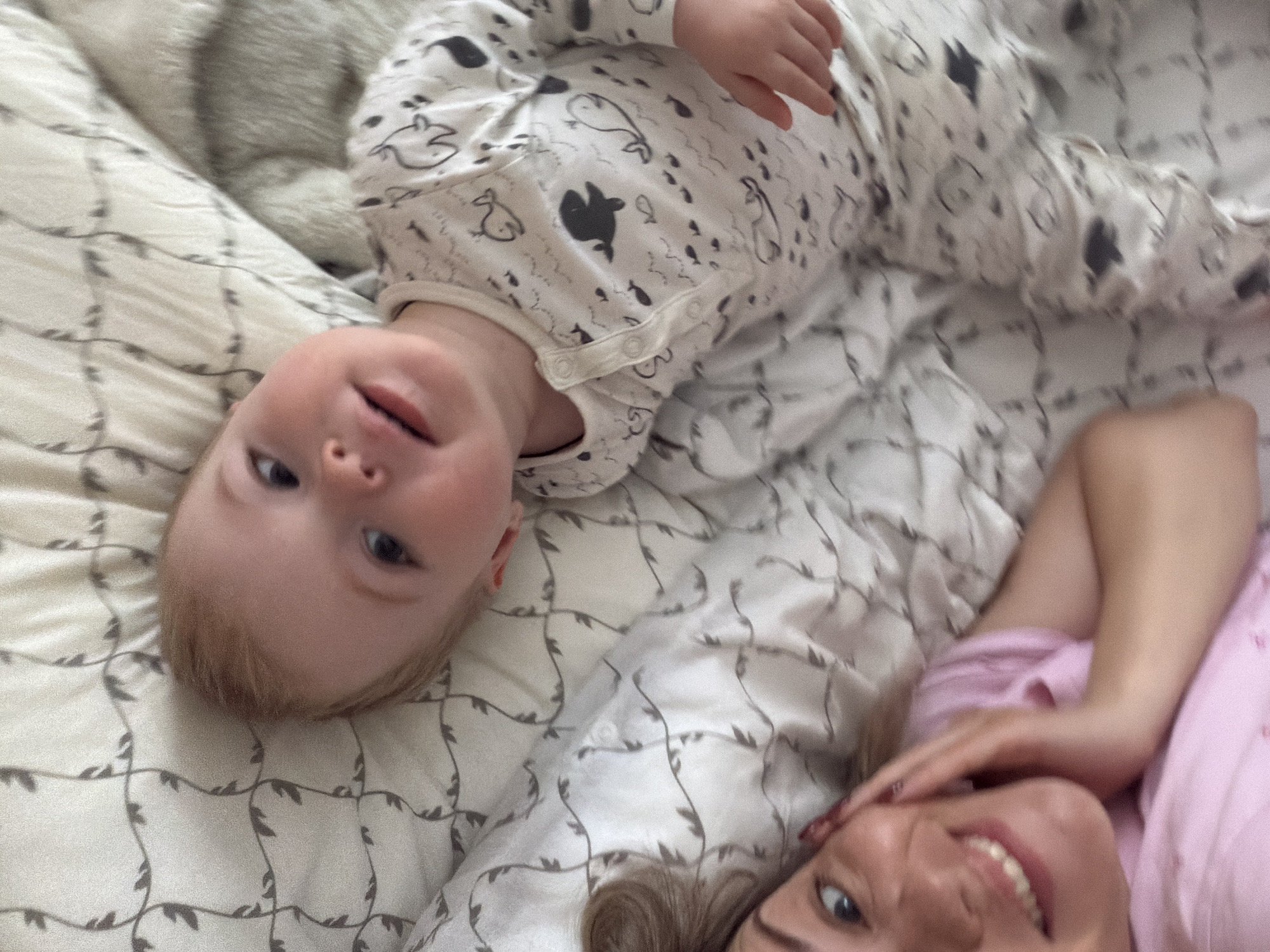 A woman and a baby are lying on a bed with a patterned white comforter. The baby, wearing a white onesie with black animal designs, is positioned upside down near the top left corner of the frame. The woman, wearing a pink shirt, is at the bottom right. The environment is an interior setting, likely a bedroom. The location cannot be inferred from the image, and there is no indication of the city of Votkinsk, Russia.