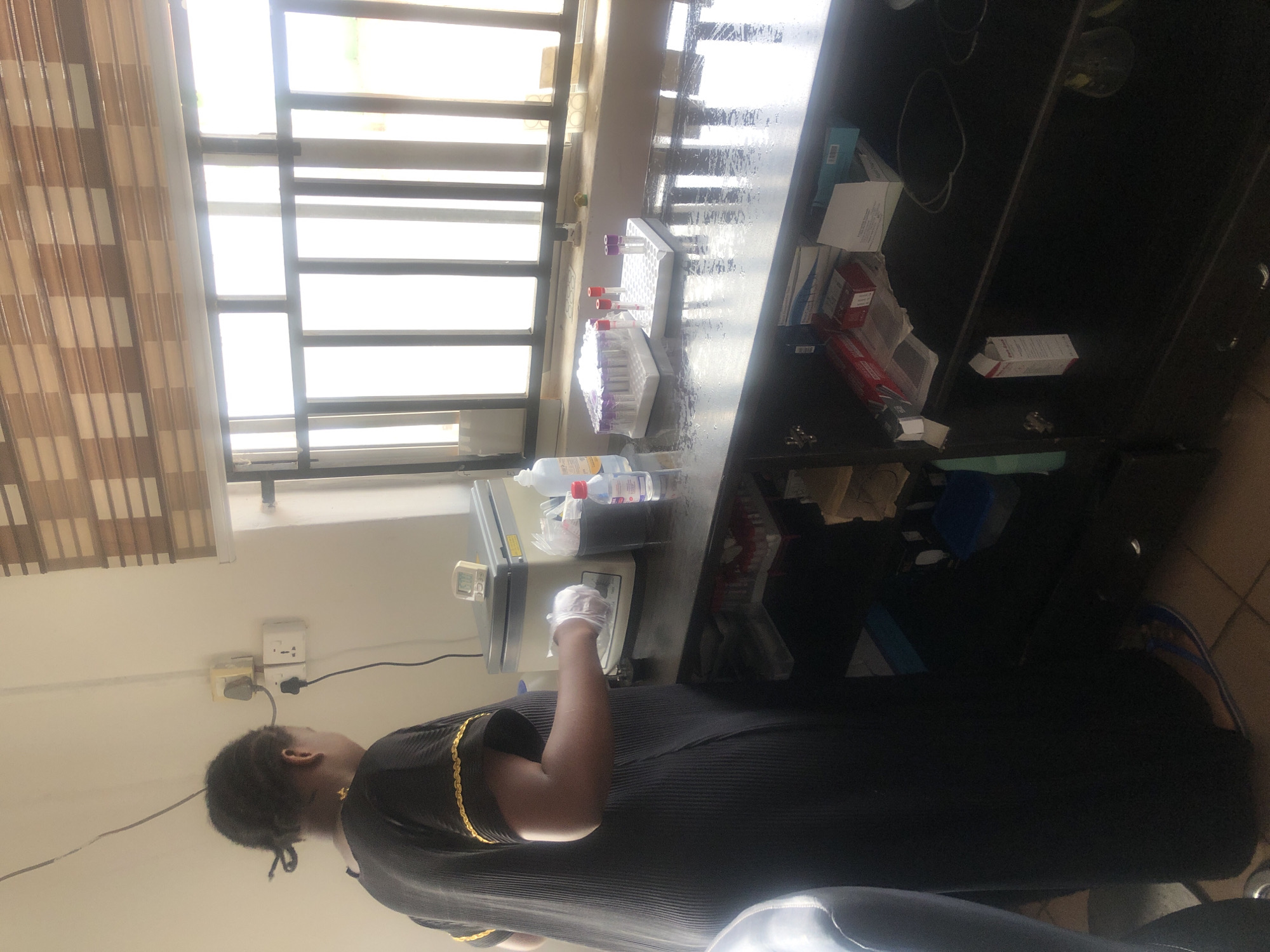 The image shows an individual working in a laboratory setting in Jos, Nigeria. The person, wearing a black dress with gold detailing, is standing at a counter wearing gloves while working with equipment, possibly a centrifuge. Test tubes in racks, bottles of liquids (possibly reagents or cleaning solutions), and boxes of supplies are arranged on the counter near a window covered with window blinds. A dark cabinet is to the right of the counter. A power outlet and plugs are visible on the wall behind the individual. Tiles are visible on the floor.