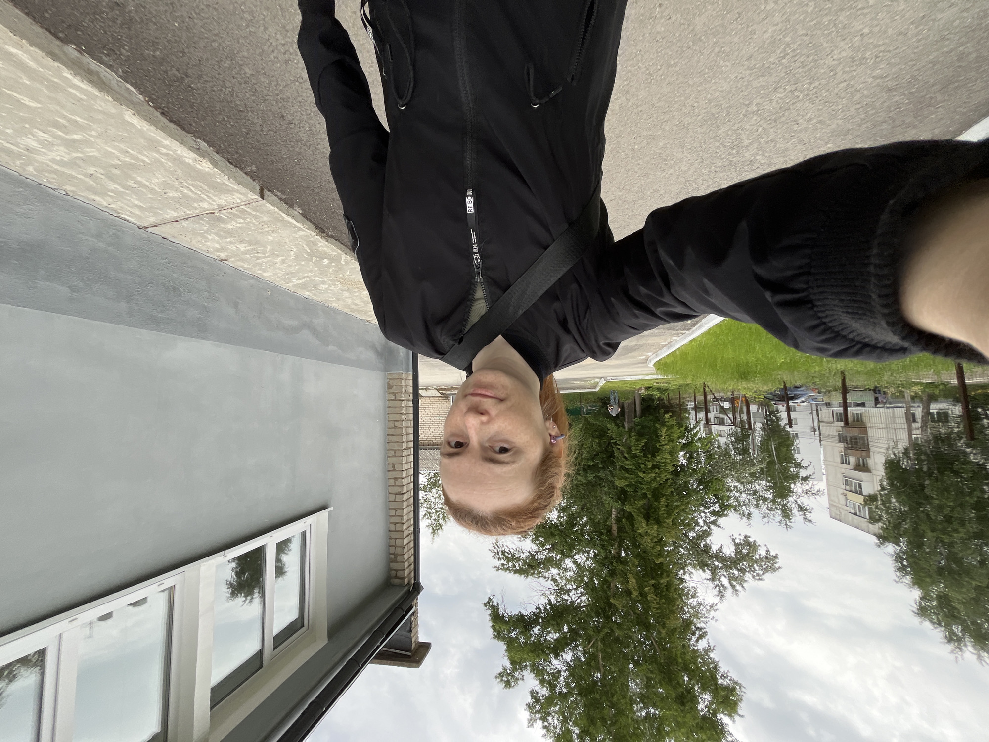 The image is a selfie taken by a person outdoors in Chaikovsky, Russia, depicting an urban environment under a cloudy sky. The person, with reddish-blonde hair and a neutral expression, is wearing a black zipped jacket. A dark strap, possibly from a bag, is visible across their chest. The zipper pull on the jacket clearly displays the text "RE-BORN" vertically.
To the left of the person is a light grey building with white-framed windows that reflect the sky and surrounding trees. A dark roof edge or gutter is also visible on this building. The ground features a mix of light-colored pavement, possibly a sidewalk, bordering a darker asphalt road.
The background includes abundant green grass and numerous trees with lush foliage, indicating either late spring or summer. Through the trees, a multi-story residential building with balconies is visible, consistent with the architecture found in Russian cities. The overcast sky suggests diffused daylight. The overall scene captures a casual, everyday moment in an urban setting.