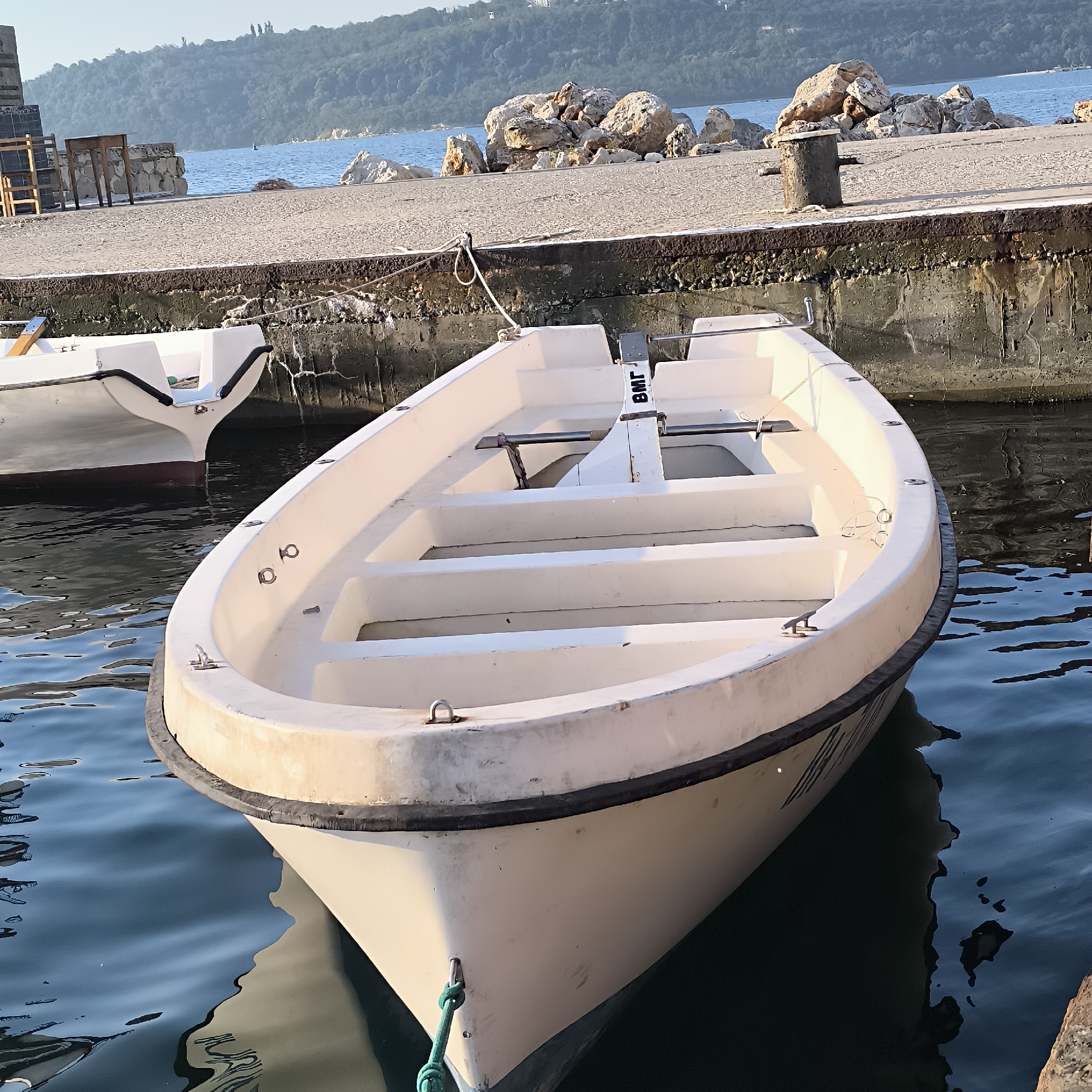 A white, open-decked boat is moored in calm, dark blue water next to a concrete pier. The boat is positioned at an angle, with its bow facing the viewer. Inside the boat, several wooden or plastic benches are visible. A black trim runs along the top edge of the boat. A green rope is tied to the bow. In the background, the pier is made of concrete and lined with a rough, textured edge. Beyond the pier, large, light-colored rocks are piled up. The water extends to a horizon where a green, tree-covered hill is visible under a clear, light blue sky. The overall scene appears to be a harbor or marina. The presence of the water, pier, and boats suggests a coastal urban environment. The specific location is a quay in the Sea Garden area of Varna, Bulgaria, likely near the yacht club or naval facilities.