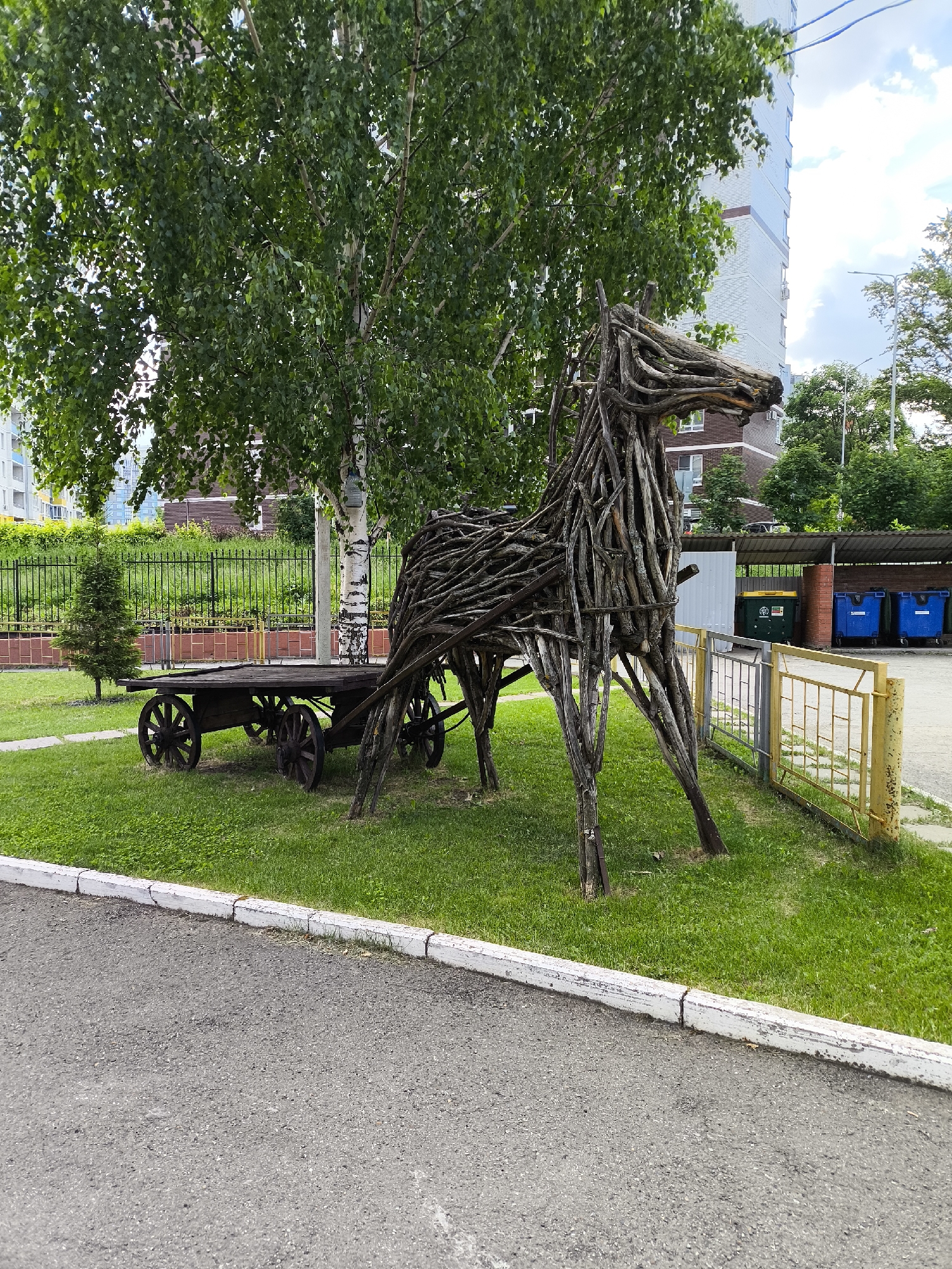 The image displays an urban green space, likely a courtyard or park area, featuring a large, stylized sculpture of a horse pulling a cart. The horse sculpture is constructed from interwoven branches and twigs, standing on four legs with a distinct head and mane, positioned on a vibrant green lawn. Behind the horse, a dark wooden cart with four large, spoked wooden wheels is attached.
A prominent birch tree with white bark and lush green leaves stands to the left of the sculpture, casting some shadow. Further left, a small evergreen sapling is visible near a black metal fence line. In the background on the left, several multi-story residential buildings with light-colored facades are partially visible through the trees.
To the right of the horse sculpture, a low yellow and silver metal fence delineates the grassy area from an asphalt surface. Beyond this fence, a covered waste collection point houses one large green industrial waste bin and two large blue industrial waste bins. In the further background on the right, another tall residential building with light-colored siding and brown lower sections is visible, along with additional trees and streetlights.
The immediate foreground consists of a grey asphalt path or road, bordered by a weathered white concrete curb that separates it from the green lawn. The sky is partly cloudy, indicating daylight. No people are present in the scene. The architecture, vegetation, and urban infrastructure depicted are characteristic of residential areas within cities such as Izhevsk, Russia.