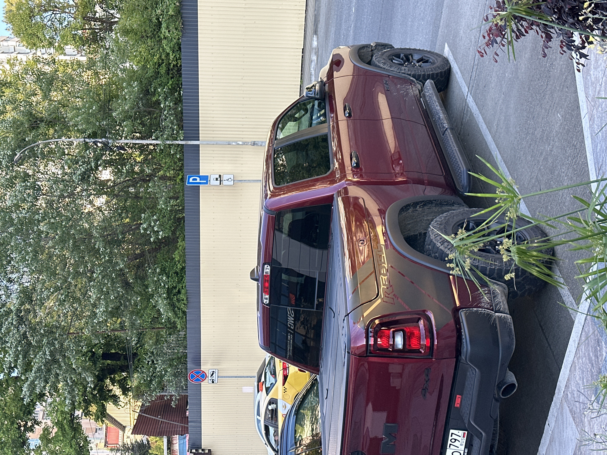 A maroon Ram 1500 Rebel pickup truck is parked in a designated parking space on a paved surface. The truck is facing right, with its rear slightly angled towards the viewer. The truck has a black tonneau cover over the bed. A license plate with the characters "797" and "RUS" is visible on the rear bumper. To the left of the truck, a yellow and white taxi is visible, partially obscured. In the background, a light-colored corrugated metal fence runs horizontally, with trees and buildings behind it. A lamppost with a blue "P" parking sign and a disabled parking symbol is also present in the background. The scene appears to be in an urban environment, likely Sochi, Russia, given the license plate format. There are no people visible in the image.