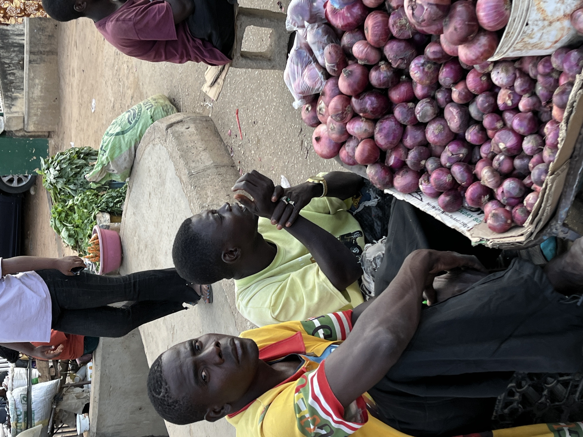 In Kabong, Nigeria, two dark-skinned men sit adjacent to a large display of red onions at a market stall. One man is eating, while the other looks at the camera. The scene also includes bags of what appear to be produce, and the legs of a person in black jeans and a white shirt are visible to the left. A car is visible in the background, along with more goods on sale.