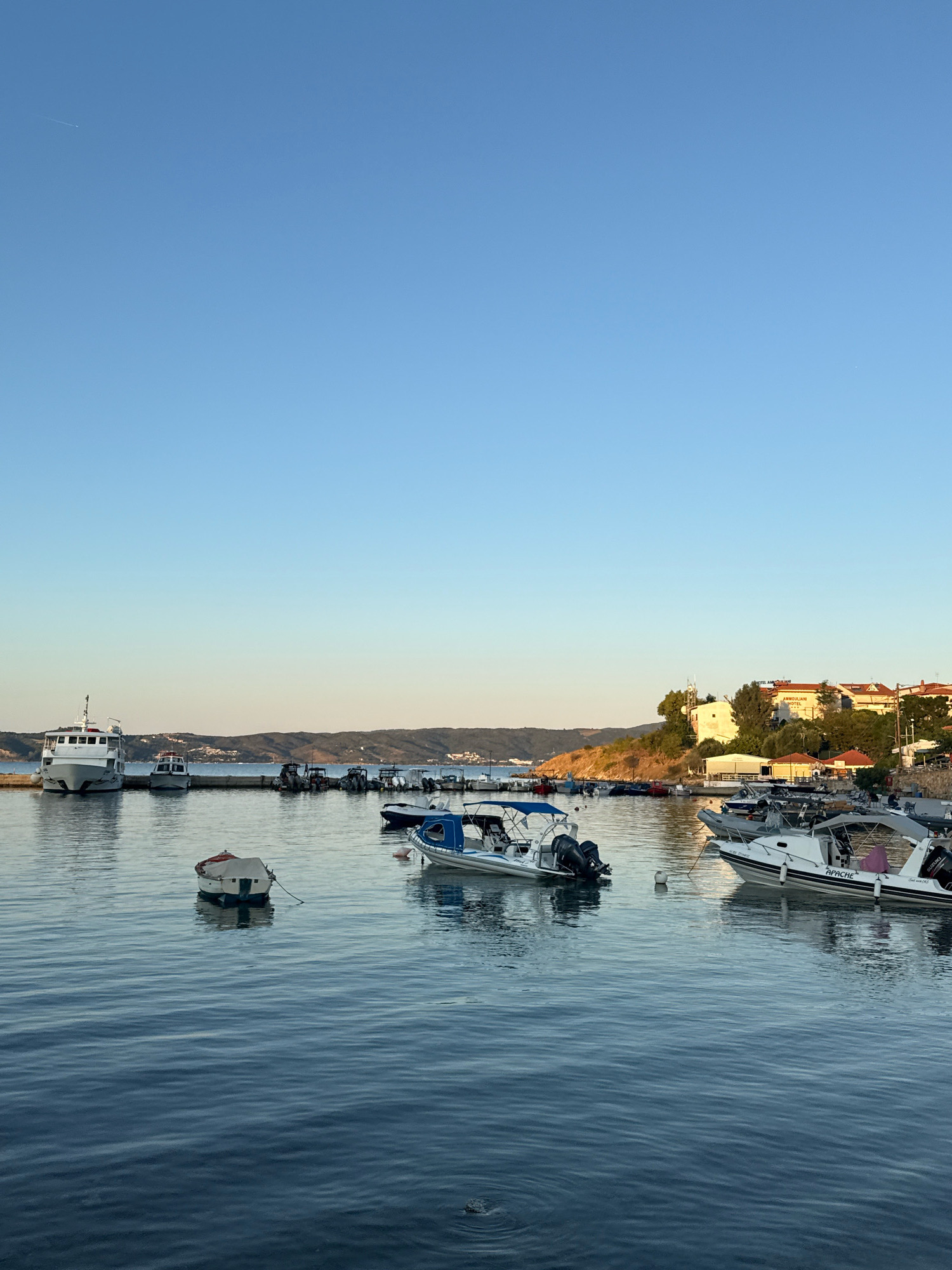 A wide, tranquil shot captures a peaceful harbor in Ammouliani, Greece, under a clear, pale blue sky. The water, a deep, calm blue, reflects the sky and the surrounding boats. Several boats are docked or moored, varying in size and type. In the foreground, a small, white boat with a blue canopy is tied to a mooring. To the left, a larger white ferry is docked. Behind the boats, a picturesque coastline rises, dotted with buildings and lush green trees, suggesting a Mediterranean setting. The overall atmosphere is serene and idyllic, bathed in the soft light of what appears to be late afternoon or early morning.