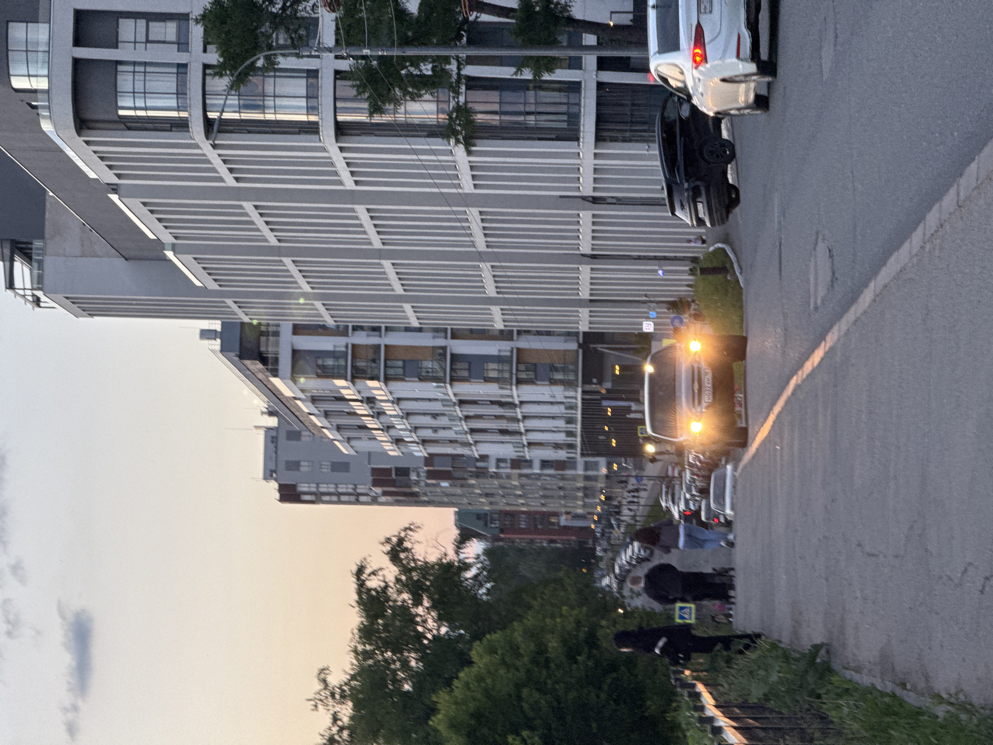 The image depicts an urban street scene during dusk or early evening in Izhevsk, Russia. On the left, several modern, multi-story residential buildings dominate the view. They feature prominent gray facades with a pattern of horizontal lines and numerous windows, suggesting contemporary architecture.
An asphalt road runs along the right side of the frame, where several cars are parked. In the foreground, a white hatchback or SUV and a dark car are visible. Further down the street, a vehicle, possibly a van or larger car, approaches or is stopped, with its bright headlights illuminating the immediate area. A partially visible license plate on this vehicle shows the numbers "700".
To the right of the road, a grassy bank slopes down, and several green trees are scattered along the street and in the foreground. Two individuals are visible on the right side, on what appears to be a sidewalk or pathway. One person is wearing dark clothing, possibly a hoodie. A small blue square sign with a white triangular pedestrian symbol is visible near them.
The sky above displays the soft hues of twilight, transitioning from a light orange-pink near the horizon to a lighter gray-blue, with a few faint clouds. The activated headlights and the sky suggest the transition from day to night. The scene captures a quiet moment in a residential area as evening descends.