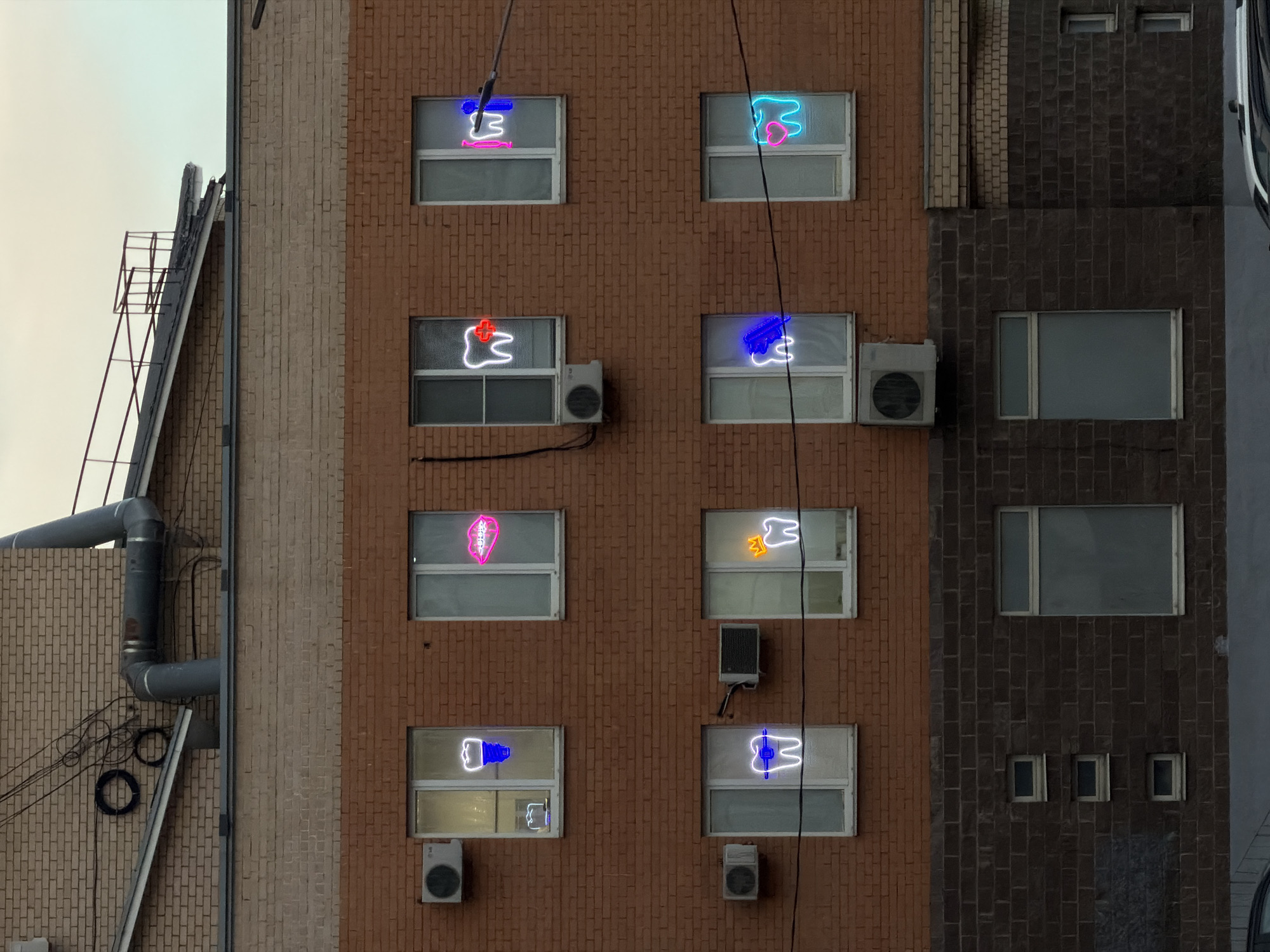 This image displays the facade of a multi-story brick building in Khimki, Russia. The upper two visible floors consist of reddish-brown brickwork, while the lower section is constructed from darker brown/grey brick. Eight distinct windows, framed in white, are arranged in two horizontal rows of four on the reddish-brown section of the building. Each window contains an illuminated neon sign, predominantly featuring dental motifs.
From left to right, the top row of neon signs depicts:
1. A white tooth with a blue implant screw base.
2. Pink lips displaying white dental braces.
3. A white tooth overlaid with a red cross or plus symbol.
4. A white tooth positioned beside a pink human silhouette holding a blue tool.
From left to right, the bottom row of neon signs shows:
1. A white tooth featuring blue dental braces.
2. A white tooth adorned with a yellow crown.
3. A white tooth with a blue stylized shape, possibly representing a tool or hand.
4. A white tooth containing a blue heart within its outline.
Several external air conditioning units are mounted on the brick facade near various windows. A large, grey ventilation pipe runs horizontally along the upper section of the building before bending upwards. Numerous black electrical cables are visible traversing the brickwork and some windows. The building's roofline, visible at the top, includes a dark, sloped section and a metal railing structure. The sky is overcast and uniformly light grey. The roof of a vehicle is partially visible at the bottom edge of the frame.