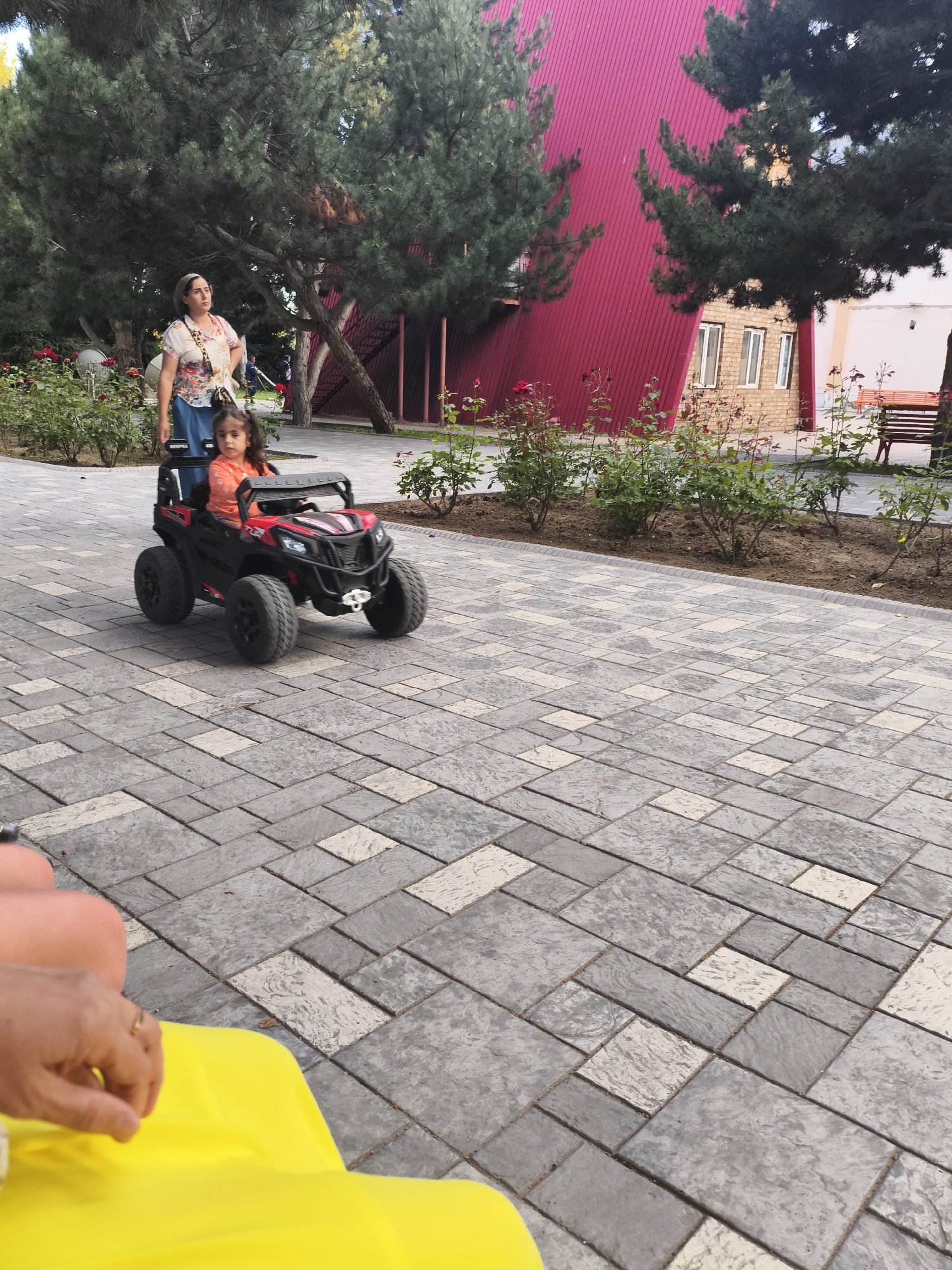 In Bayet, Kyrgyzstan, a young girl in an orange shirt drives a red toy car on a paved pathway. Behind her, a woman in a floral shirt and blue skirt stands near a rose bush. The background features trees, a building with a red corrugated metal facade, and a brick structure. The pathway is made of gray and cream colored rectangular pavers. In the foreground, a person's hand with a ring is visible resting on a bright yellow fabric.