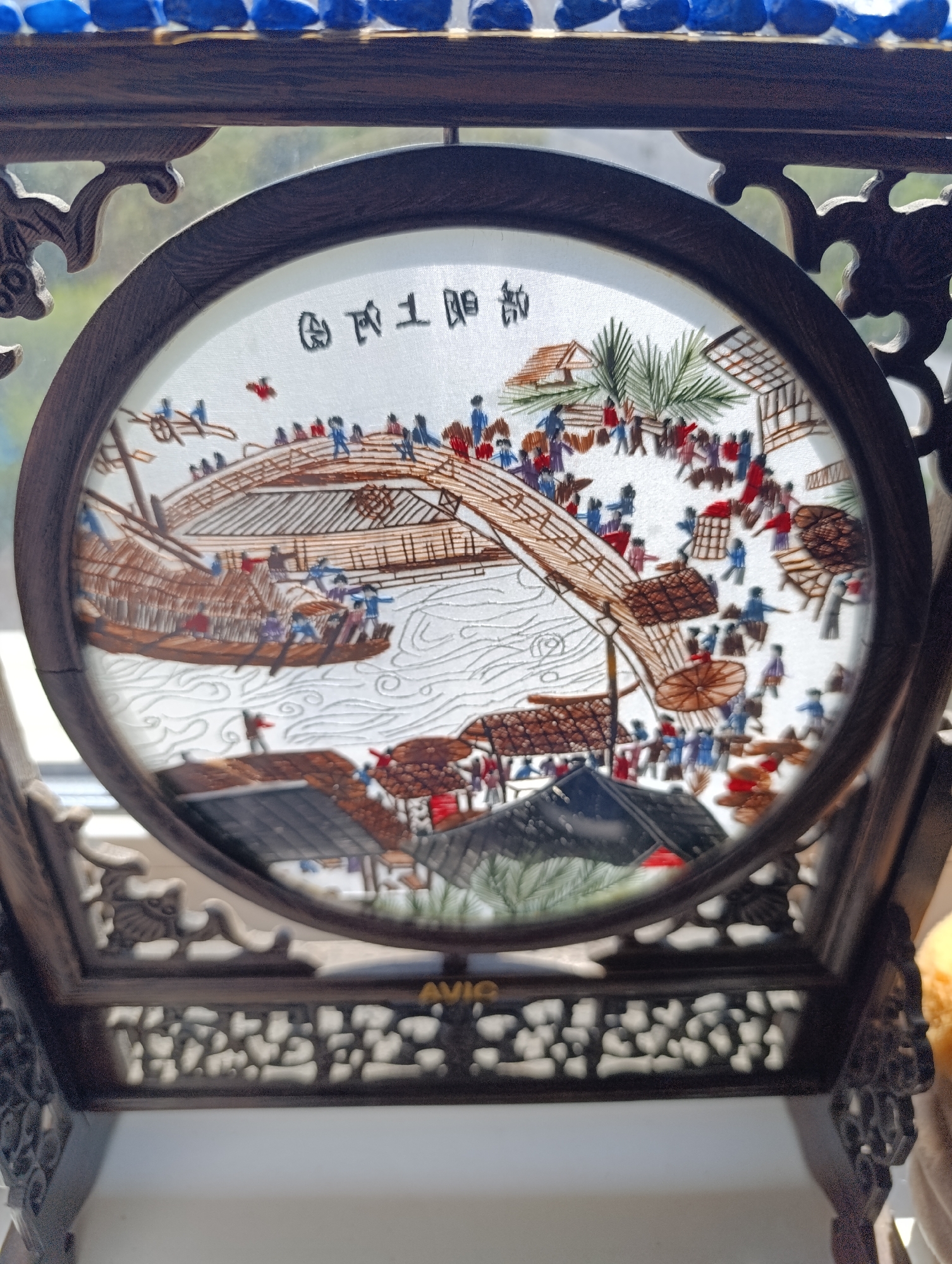 An embroidered circular artwork is displayed within a dark, ornate wooden frame. The artwork depicts a bustling riverside scene with a bridge, a boat, and many small figures. The scene is rendered in thread on a white background, creating a textured and colorful image. The frame itself is intricately carved with floral or possibly mythical motifs, and it rests on a carved wooden stand. The background behind the artwork is out of focus, suggesting it is placed near a window or in a well-lit area. The overall impression is of a traditional Chinese or East Asian decorative piece.