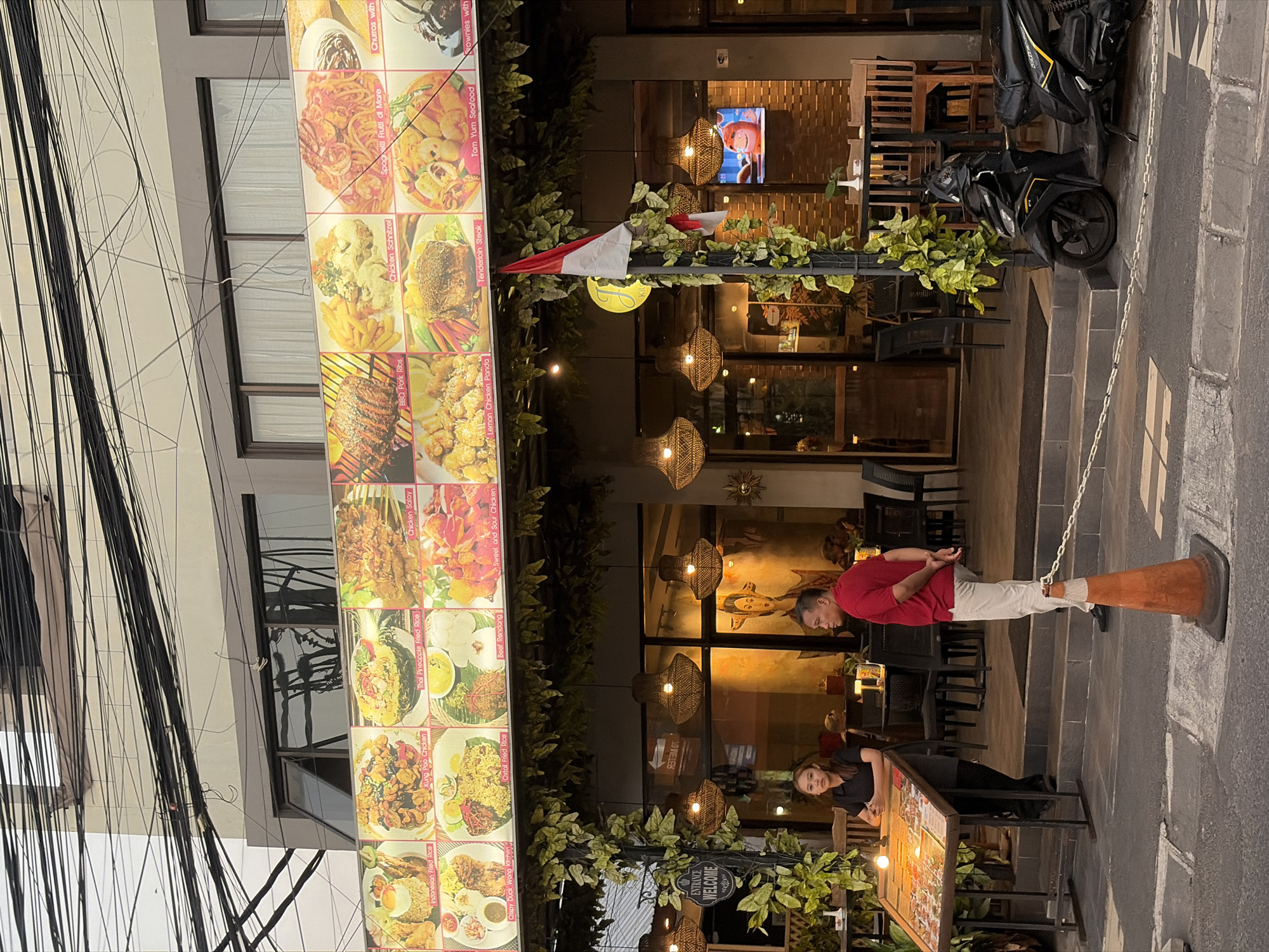The image captures a street scene in Kuta, Indonesia, featuring a restaurant with an outdoor dining area and a prominent menu board. A young woman in a black uniform stands behind the menu board, appearing to be an employee. A man in a red polo shirt and white pants stands to the right of the menu, facing away from the camera and towards the restaurant entrance. The restaurant's facade displays a large illuminated menu with images of various dishes. Hanging plants and decorative lights adorn the entryway. A motorcycle is parked on the sidewalk in front of the establishment. The street is paved, and overhead electrical wires are visible against the sky, characteristic of urban infrastructure in the region. An orange traffic cone is positioned on the road. The overall atmosphere suggests an active commercial area in Kuta.
