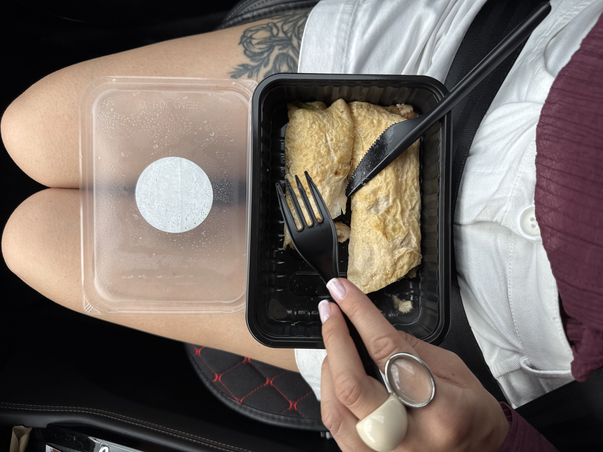 A person's legs are visible in the frame, one leg crossed over the other, wearing what appears to be a white skirt and a burgundy top. On the lap is a black plastic takeout container holding two portions of what looks like a savory crepe or omelet, with a fork and knife positioned above it. To the left of the food container is an empty clear plastic lid, covered in condensation droplets, with a circular sticker in the center. A tattoo of floral imagery is visible on the upper part of the left leg. The person is wearing a distinctive ring on their right hand, featuring a large, oval, clear stone and a bulbous cream-colored accent. The background appears to be the interior of a car, indicated by the black seat upholstery with a diamond-stitched pattern. The overall impression is of a casual meal being consumed on the go. There is no visible text other than the faint imprint on the plastic lid, which is upside down and illegible.