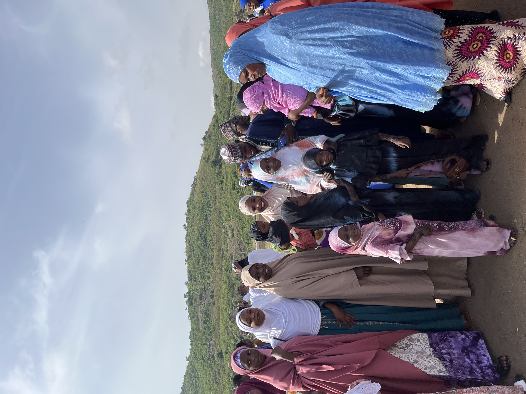 The image shows a group of people, mostly women and children, standing together. Many of the women are wearing hijabs in various colors, including blue, pink, white, and brown. They appear to be standing outdoors on a dirt surface, with a hillside visible in the background. The sky is light blue with some clouds. The location is the city of Naraguta Hausawa, Nigeria.