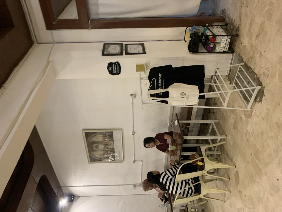 The image shows the interior of a cafe in Probolinggo, Indonesia, located at coordinates -7.751 latitude and 113.213 longitude. Two women are seated at a table in the foreground, talking. The woman on the left is wearing a black and white striped shirt and the woman on the right is wearing a red shirt. A third woman, wearing a light-colored hijab, is seated at a table in the background. The cafe has white walls, a brown wooden ceiling, and a tiled floor. There is a painting on the wall behind the table where the women are seated. There is a white rack holding a black t-shirt and a white tote bag to the right of the women. A white bench with a wooden top is in the background. There is a small cart with various objects, including a bottle of cleaner and a roll of paper towels, next to the rack. The image was likely taken during the day, as the lighting is bright and there are no shadows.