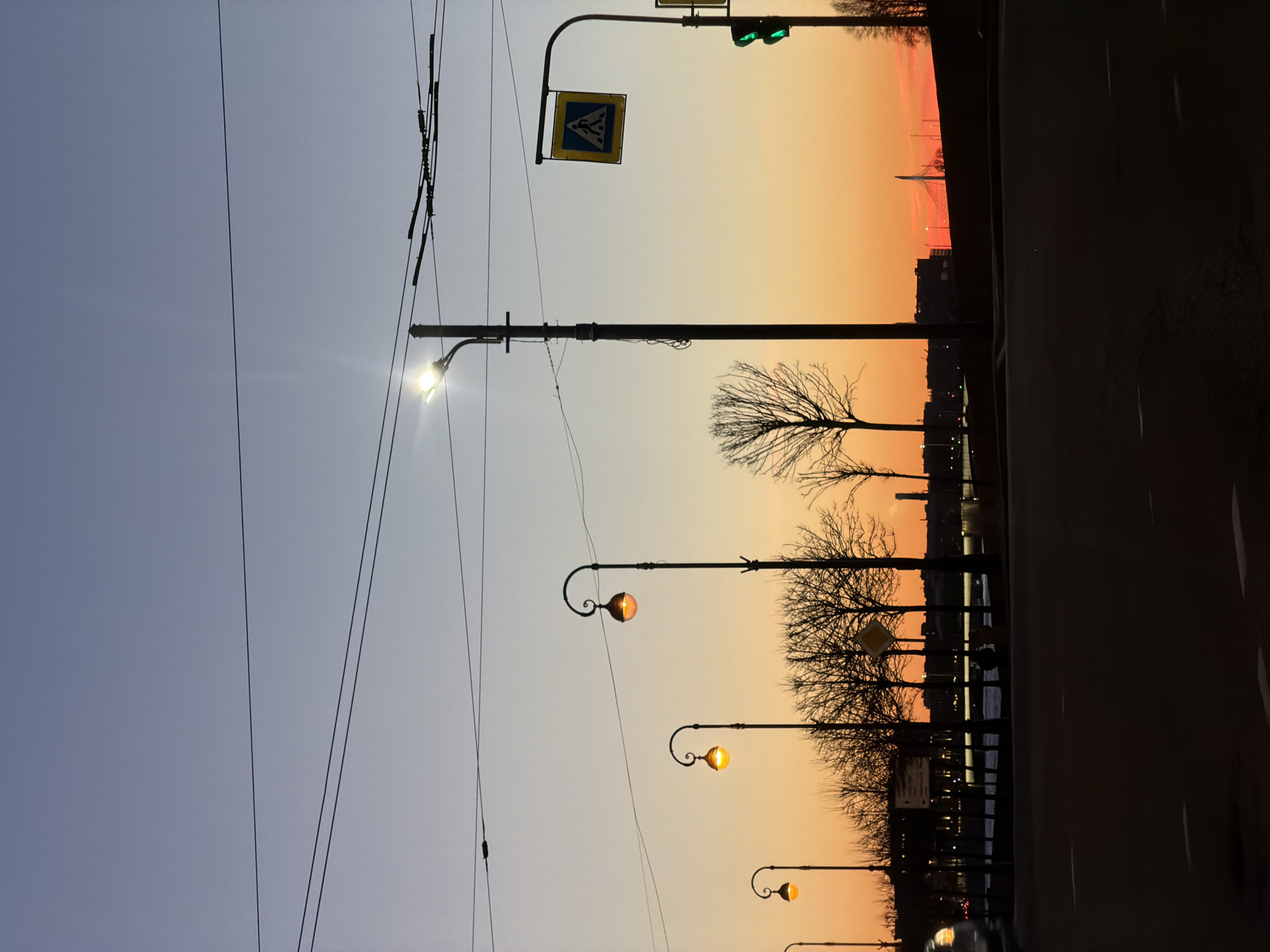 Here is a detailed description of the image:
**Scene Overview:**
The photograph depicts an urban landscape at dawn or dusk, likely alongside a river or canal, in Saint Petersburg, Russia. The sky transitions from a pale blue to warm orange hues. The lighting suggests either sunrise or sunset.
**Objects and Subjects:**
* **Street Lights:** A series of street lights line the embankment or street. These are of an older style with ornate curved arms and globe-shaped fixtures. Some are illuminated.
* **Electrical Wires:** A web of electrical wires and trolleybus power lines stretches across the sky.
* **Traffic Signal:** A traffic light is visible, with a green light illuminated.
* **Pedestrian Crossing Sign:** A pedestrian crossing sign is visible.
* **Trees:** Silhouetted trees without leaves are planted along the water’s edge.
* **Water:** Water is visible in the distance with a row of lights along the opposite bank.
* **Bridge:** In the distance, silhouetted bridge architecture is visible against the skyline. This is likely the Cable Bridge (or Bolshoy Obukhovsky Bridge).
* **Buildings:** The skyline is punctuated by building silhouettes.
* **Road:** A road takes up the right side of the image, the closest section of the road obscured by shadow.
**Location Details:**
* The scene is consistent with an embankment along the Neva River or a major canal in Saint Petersburg.
* The presence of trolleybus power lines is common in Saint Petersburg.
**Actions and Interactions:**
* It's a quiet moment, with little to no activity visibly occurring.
* The street lights illuminate the area.
* The traffic signal is functioning.
**Environment:**
* The environment suggests it is early morning or late evening in Saint Petersburg.
* The color temperature of the sky creates a warm and peaceful atmosphere.