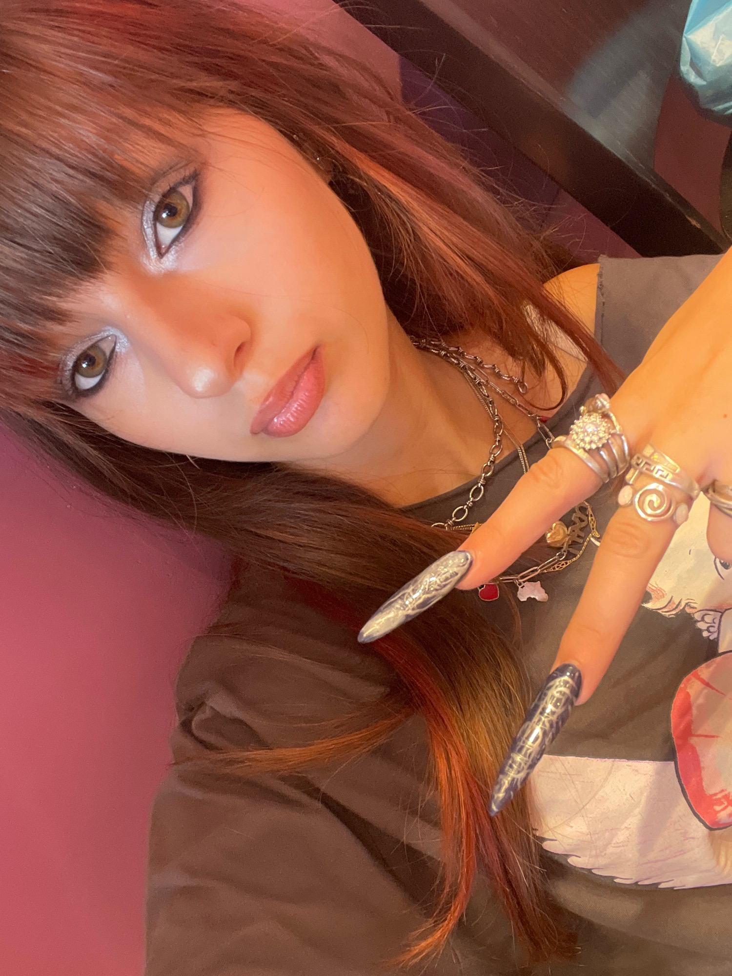 In Raanana, Israel, a young woman with reddish-brown hair and bangs is taking a selfie. She is wearing silver eyeshadow and a grey t-shirt. Her nails are long and painted with a metallic silver and blue design. She is adorned with several silver rings on her fingers and a silver chain necklace. The background is a pink wall, and a dark wooden surface is visible to her right. The overall lighting is warm, suggesting indoor lighting conditions.